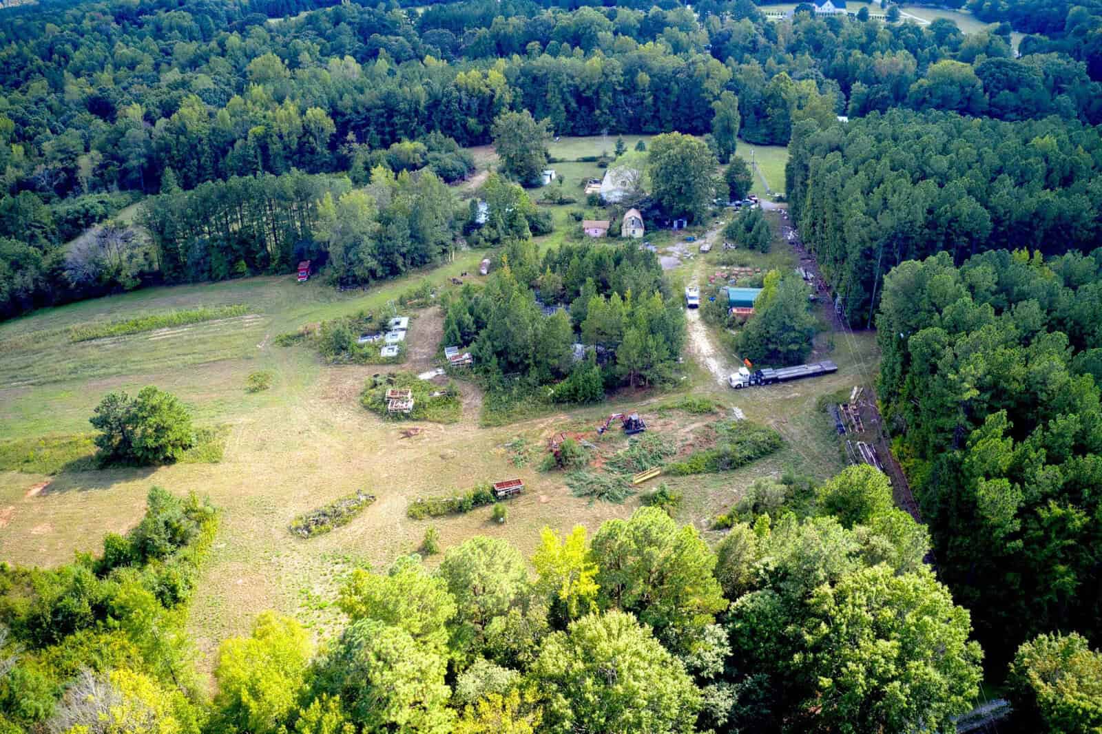 Aerial view of a rural area with scattered trees, open grassy fields, some small buildings, vehicles, equipment, and forested areas surrounding the property.
