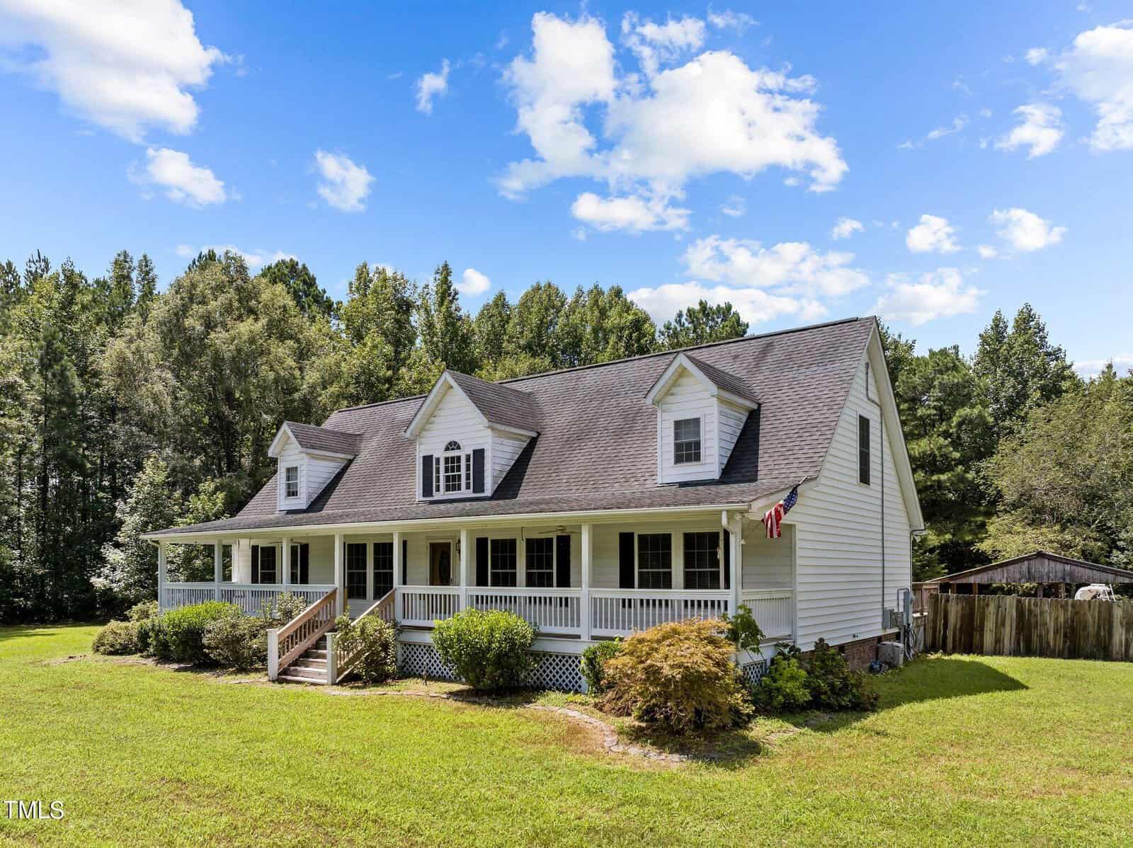 A white two-story house with a gabled roof stands in a grassy yard, surrounded by trees. It features a wide front porch with an American flag. The sky is blue with scattered clouds.