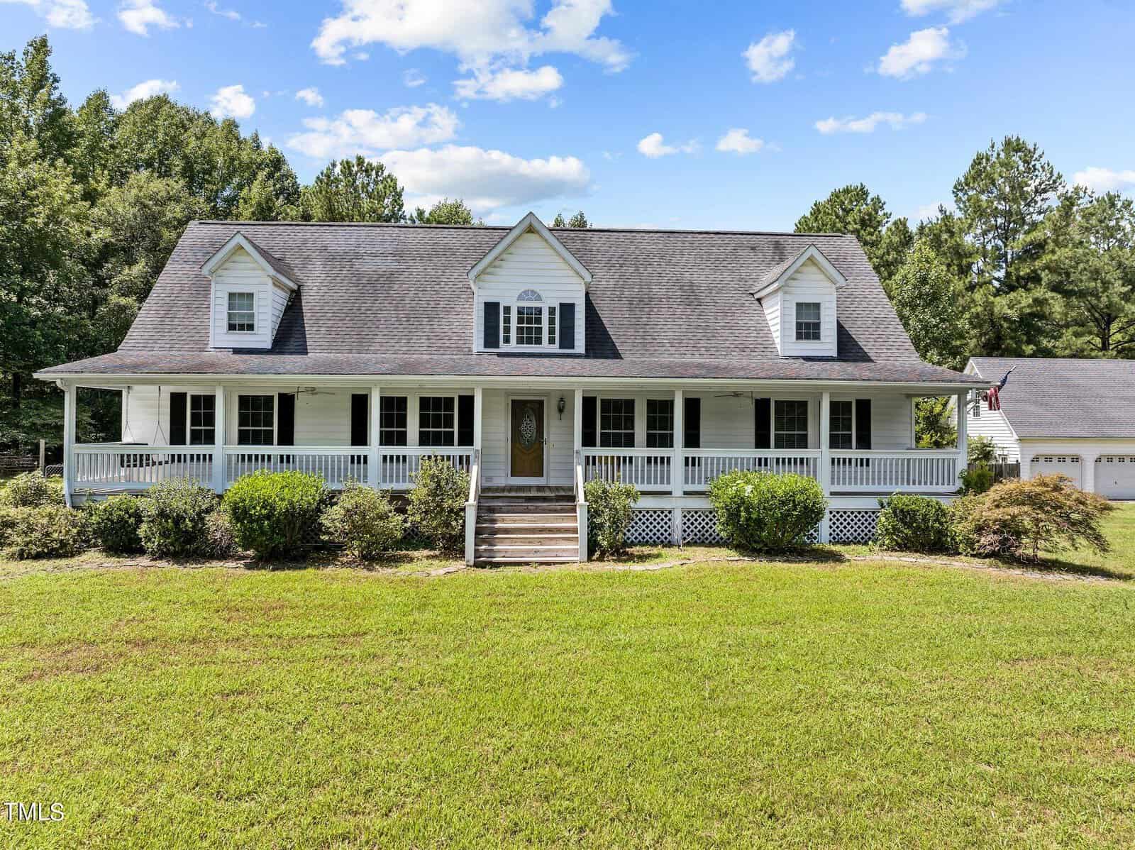Located at 5126 Misty Lane, this charming two-story Oxford-style house boasts a large front porch and three dormer windows. The white exterior contrasts beautifully with the dark gray roof. Surrounded by lush green grass and bushes, it sits under a partly cloudy sky with trees in the background.