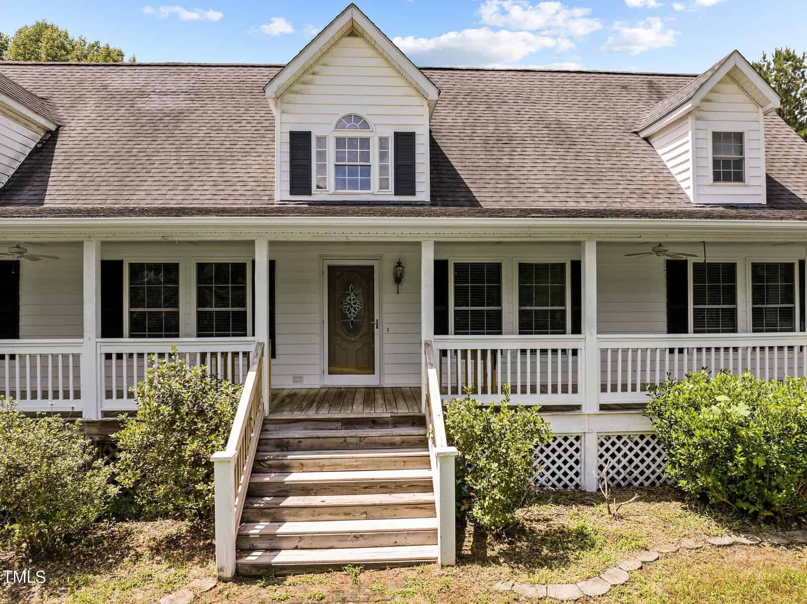A charming white house with a wraparound porch, black shutters, and a gabled roof. Two ceiling fans are on the porch, and steps lead up to the entrance. Bushes line the front, and the sky is partly cloudy.