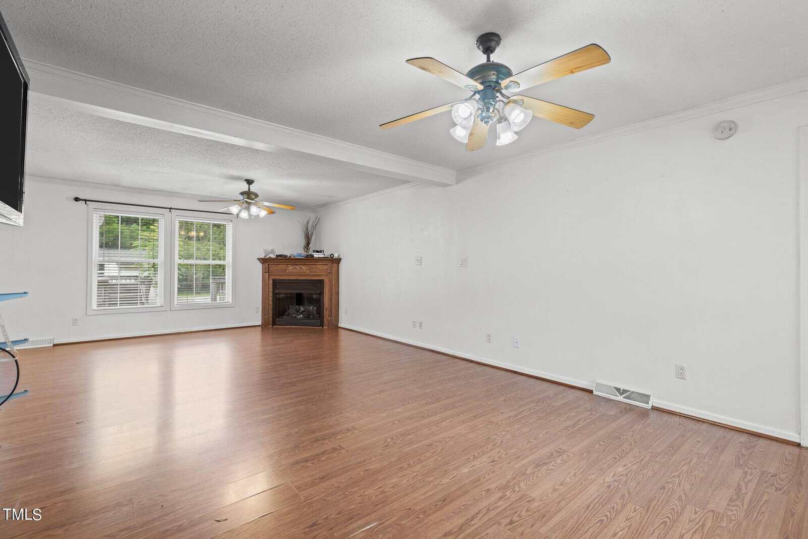Spacious living room with wooden flooring, two ceiling fans with lights, a fireplace with a mantel, and large windows allowing natural light. Walls are painted white, and room is empty of furniture.