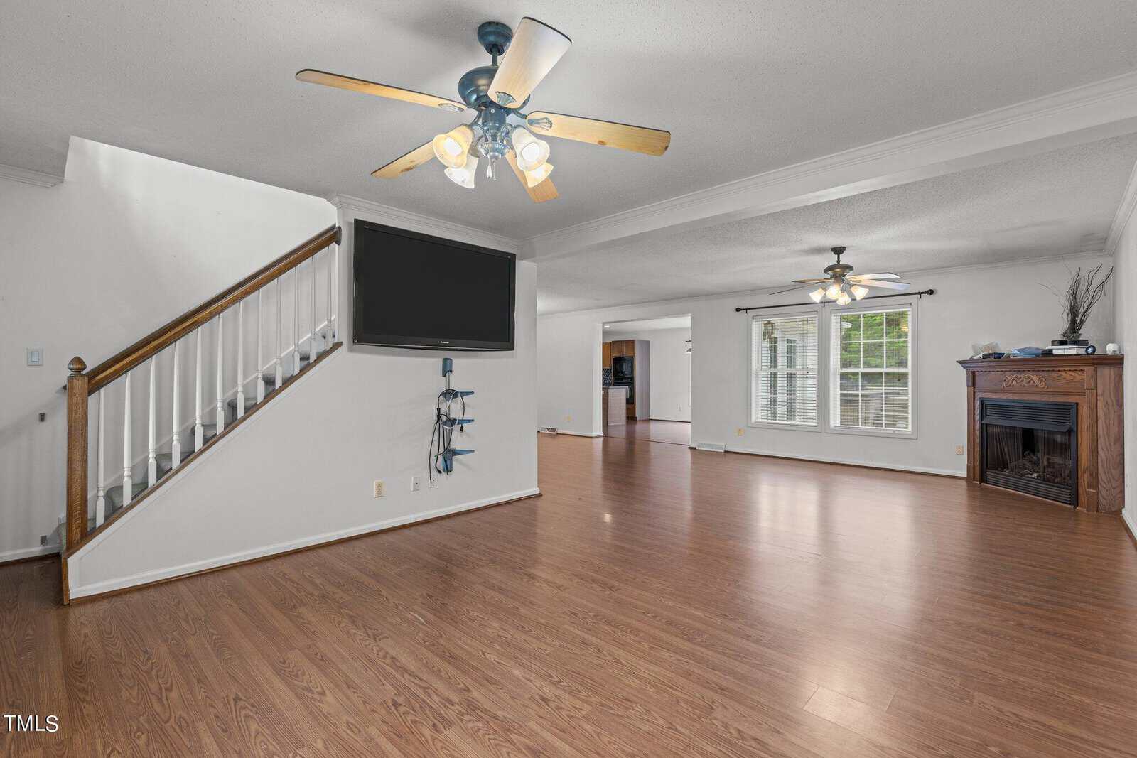 Spacious living room with wooden floors, a ceiling fan, and a wall-mounted TV. A staircase with white railings leads upstairs. Windows let in natural light, and a fireplace is on the right. An open doorway leads to the kitchen.