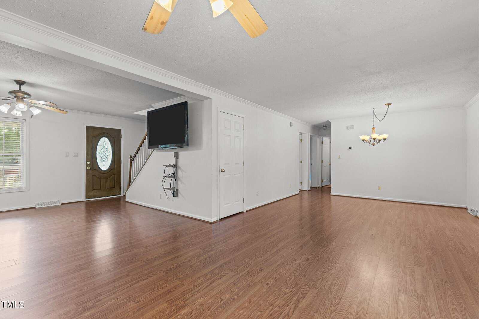 Spacious living room with hardwood floors, white walls, and a ceiling fan. A mounted flat-screen TV is on one wall. A staircase with a wooden handrail leads upstairs. A chandelier hangs in the dining area beside a window and a wooden front door.