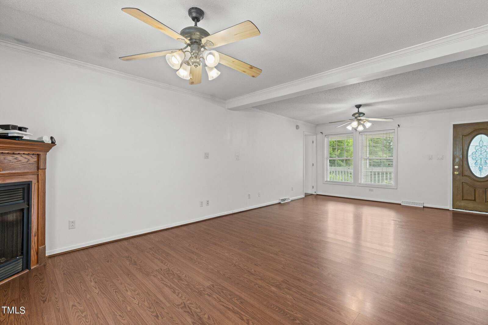 Spacious empty living room with wood flooring, white walls, and two ceiling fans with lights. A fireplace is on the left, and large windows alongside a wooden door let in natural light.