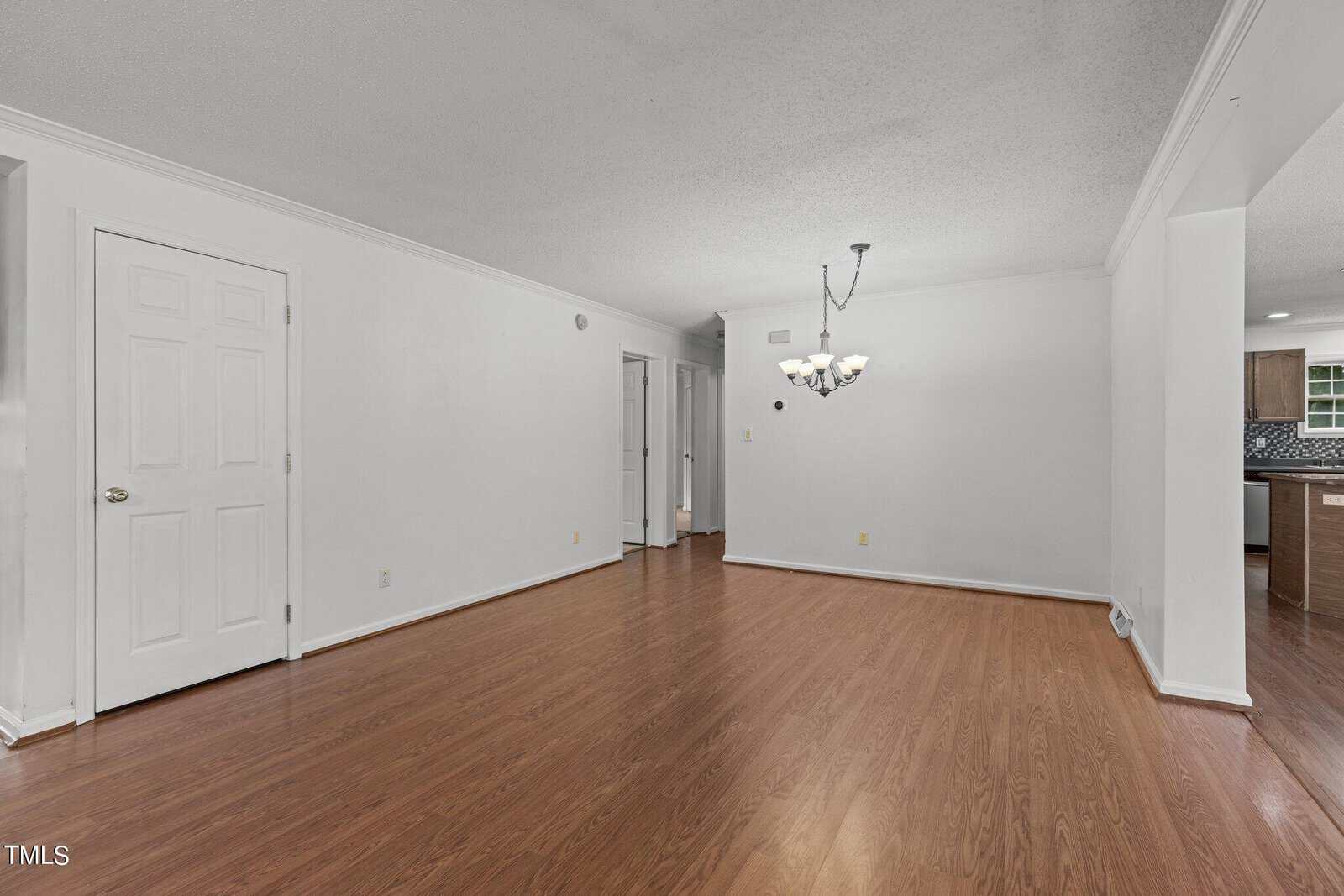 An empty room with light hardwood flooring and white walls. A ceiling light fixture hangs in the center. A closed white door is on the left, and the kitchen with cabinets and a counter is visible on the right.