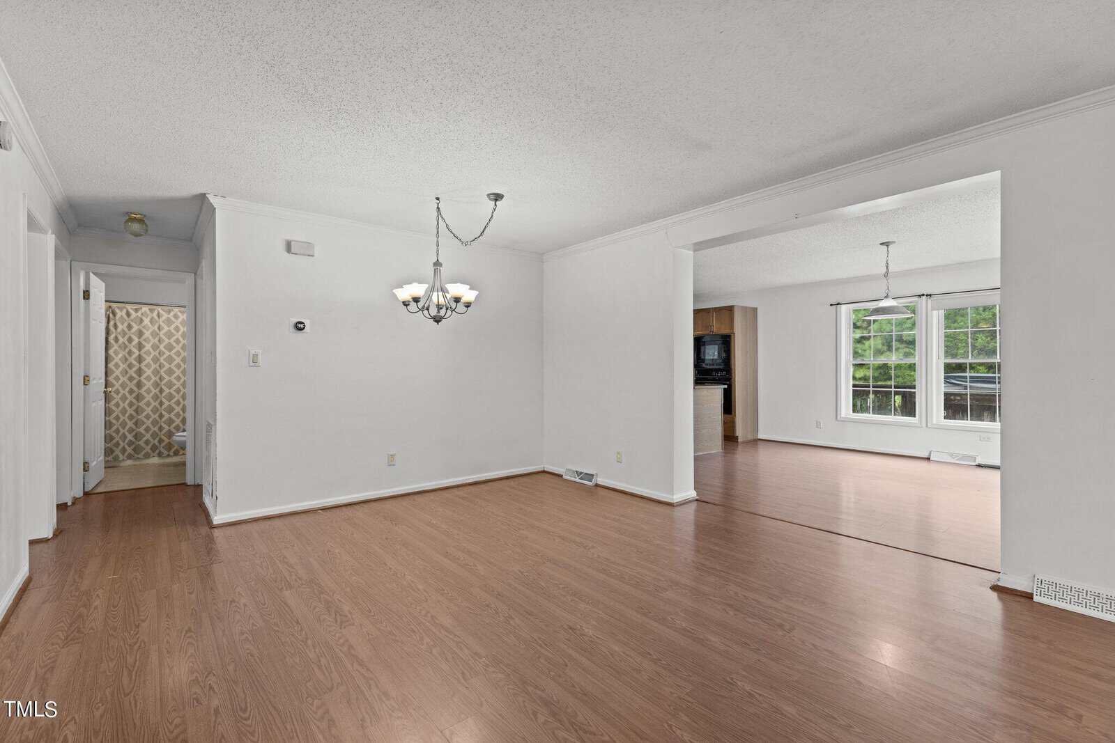 A spacious, empty living room with light wood floors and white walls. A chandelier hangs from the ceiling. A doorway leads to another room with large windows providing natural light. A hallway and patterned curtain are visible.