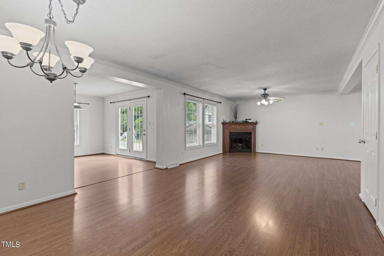 A spacious, empty living room with wooden flooring and white walls. It features a ceiling fan with lights, a fireplace, and large windows with curtains. An adjoining room with similar flooring and French doors is visible. A chandelier hangs from the ceiling.