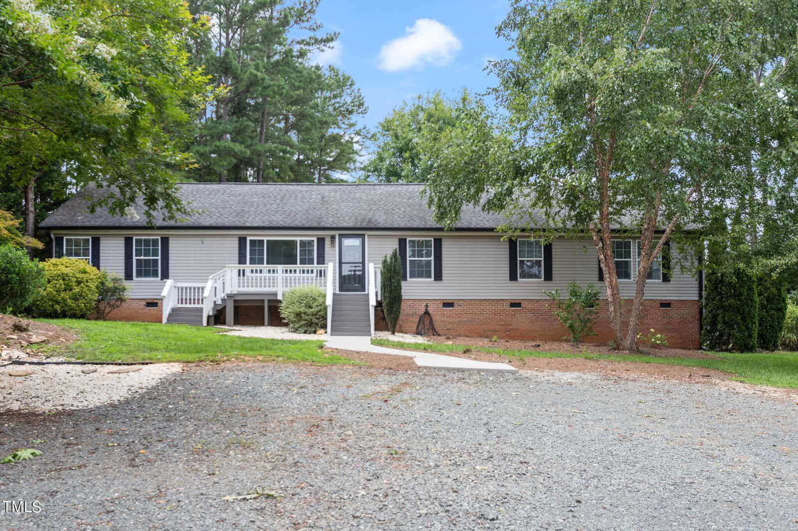 Nestled in the serene setting of Franklinton, this single-story home at 1531 Wayside Farm Road boasts a gray exterior and black roof, surrounded by lush trees and shrubs. A charming white front porch welcomes you, with a gravel driveway and patch of grass under the clear blue sky.