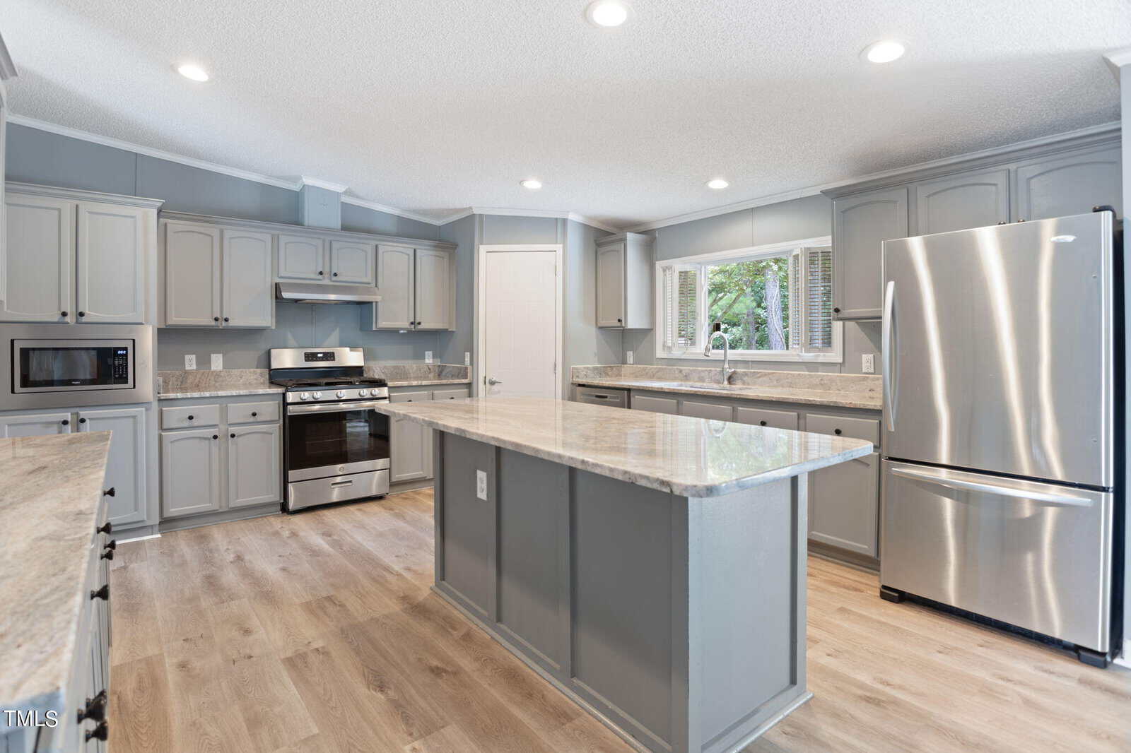 A modern kitchen with gray cabinets, a large island with marble countertops, a stainless steel refrigerator, an oven, and a microwave. The floor is wooden, and natural light comes through a window above the sink.