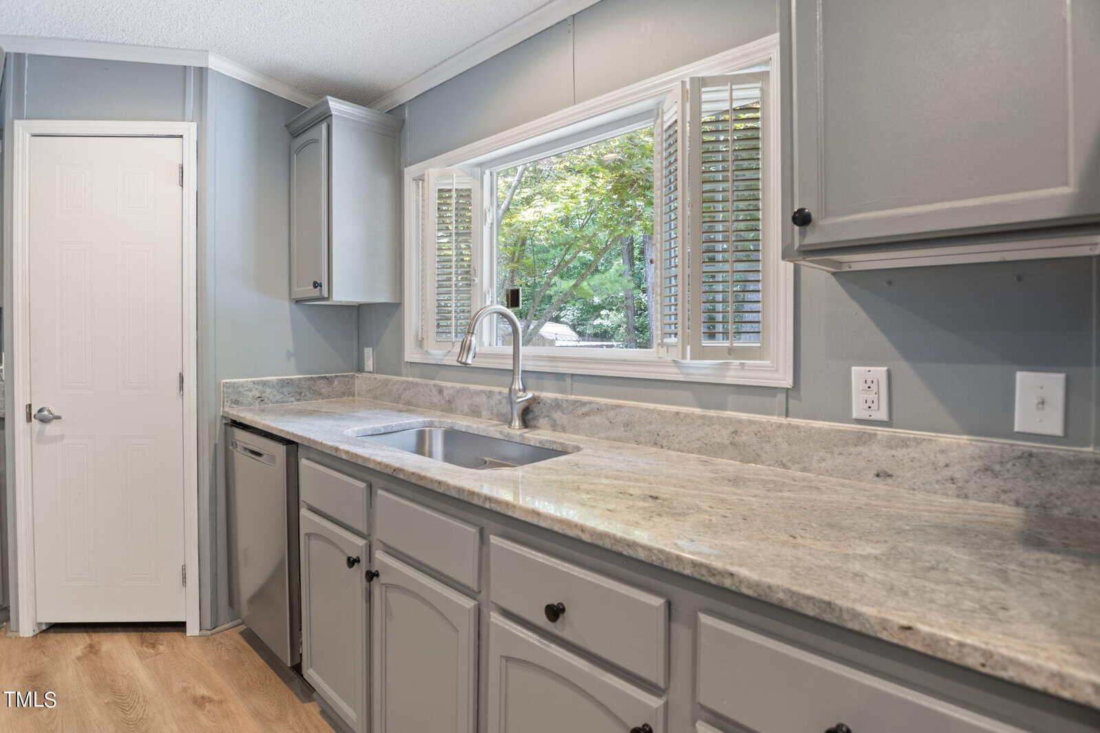 A modern kitchen with light gray cabinets, light granite countertops, and a stainless steel sink under a large window with blinds. The walls are a soft blue-gray, and the floor is light wood. A white door is on the left.