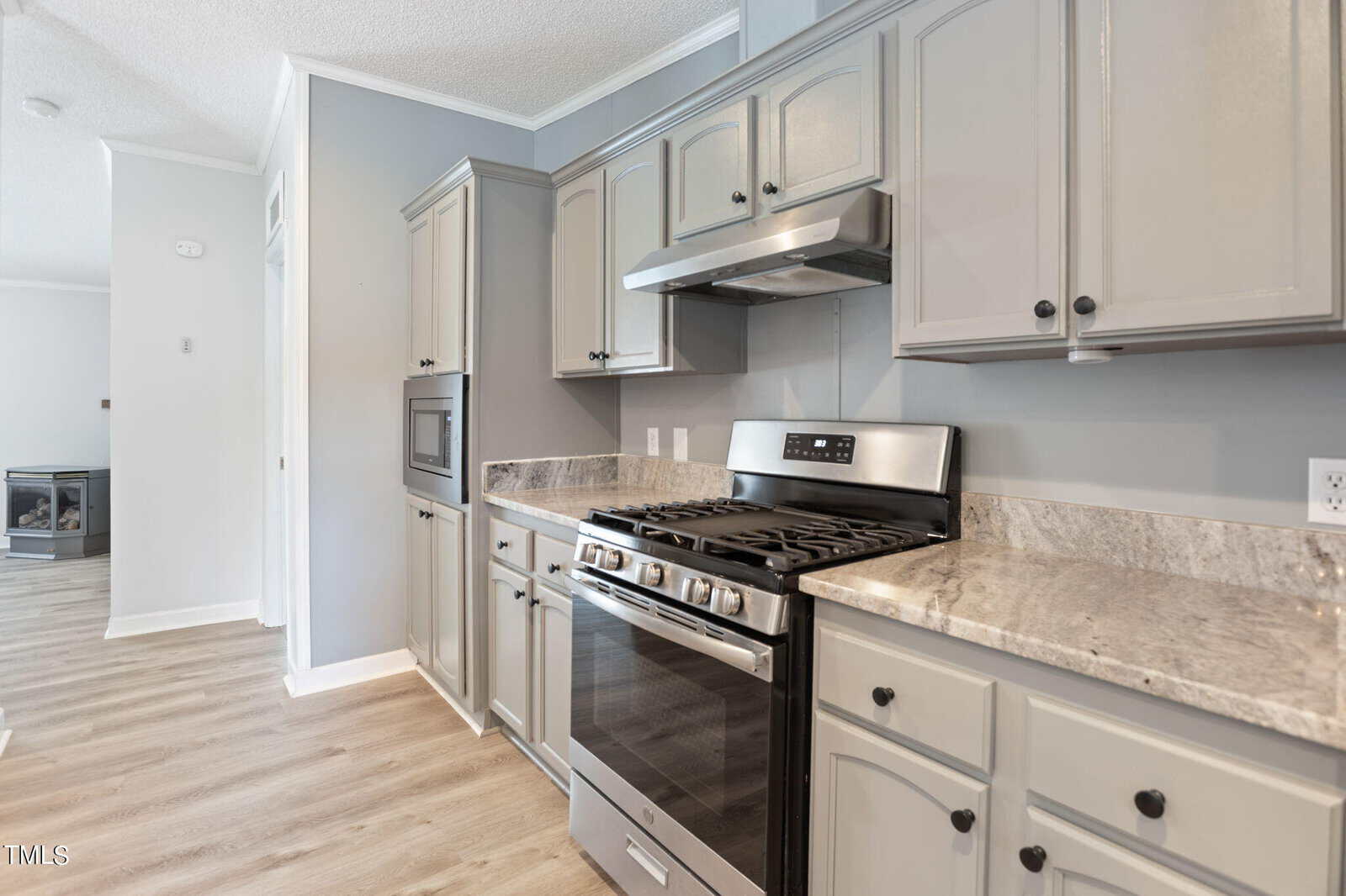 Modern kitchen with gray cabinets and marble countertops. Stainless steel appliances include a stove with oven and a range hood. Light wood flooring adds warmth, and a microwave is built into the cabinetry.