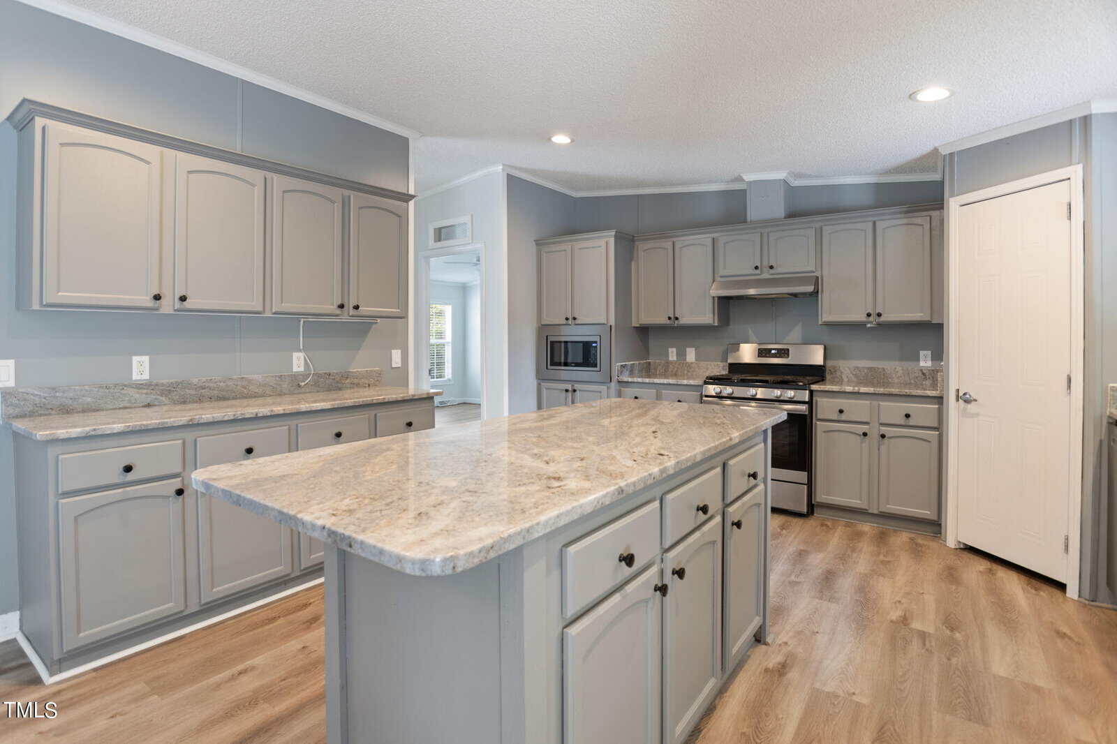A modern kitchen with gray cabinets, a marble countertop island, stainless steel appliances, and wooden flooring. The space is well-lit with recessed lighting and has a neutral color palette.