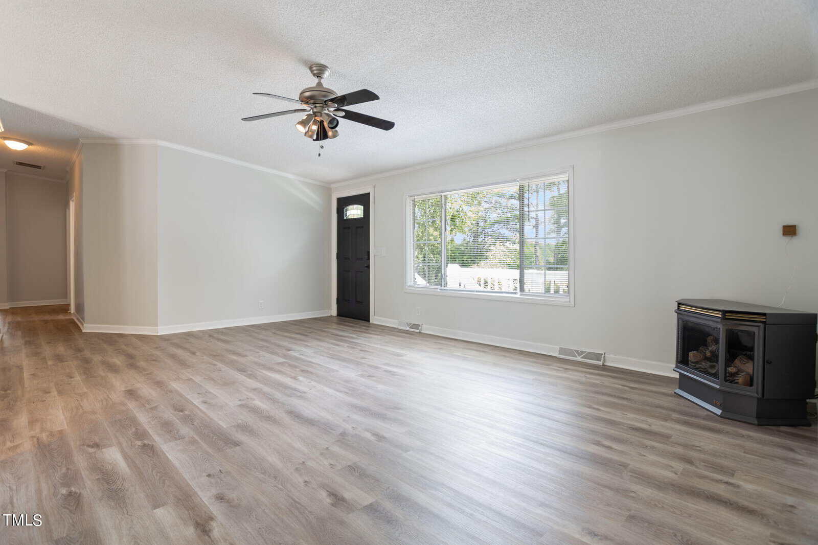 A spacious living room with light-colored wood flooring and white walls. A black ceiling fan hangs from the ceiling. A large window illuminates the room, and a fireplace sits in one corner. A hallway leads to other parts of the house.