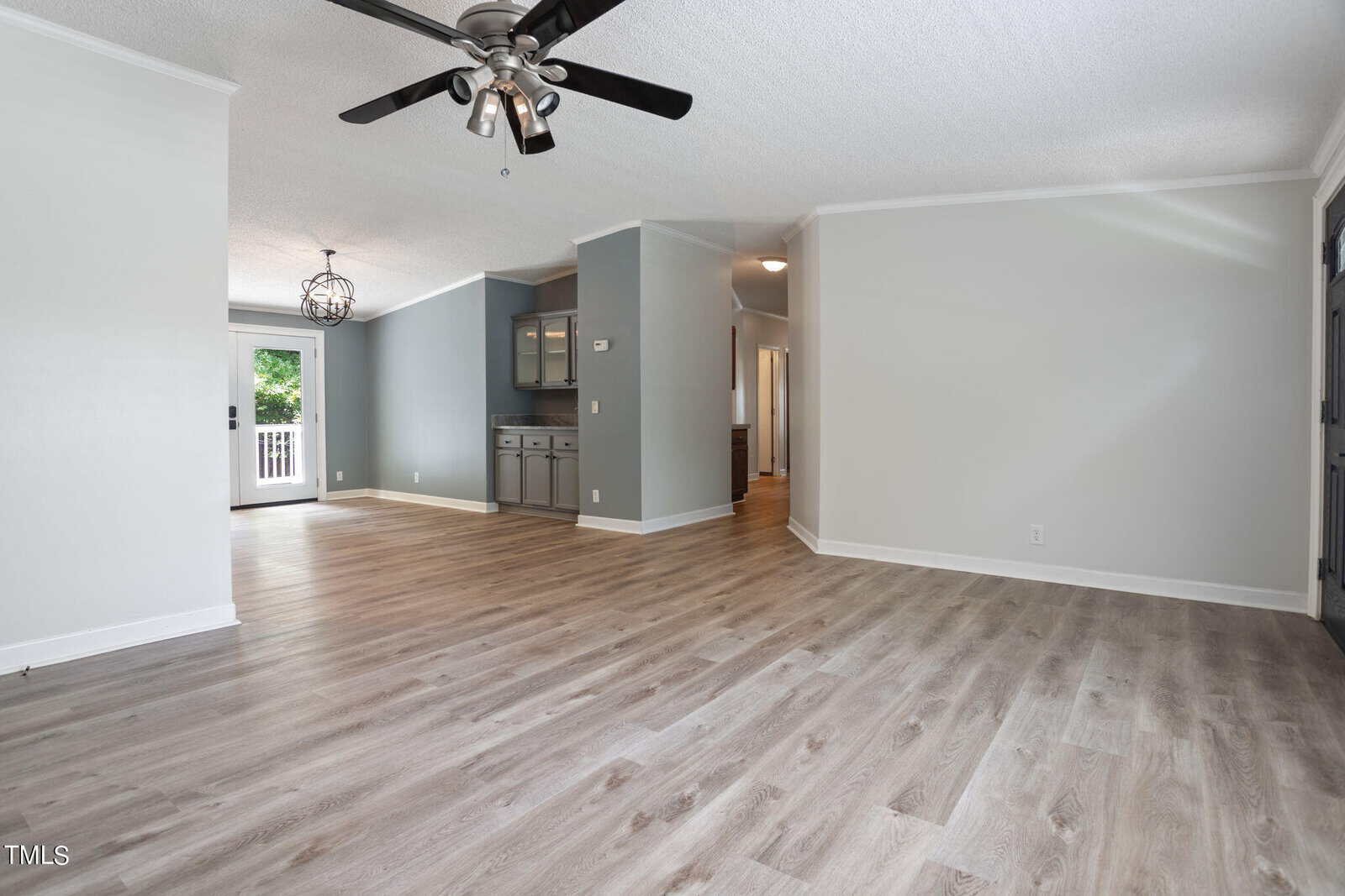 Spacious living area with light wood flooring and gray walls. Ceiling fan with black blades above. In the background, there's an open kitchen with gray cabinets and a chandelier, leading to a deck via glass doors. Natural light fills the room.