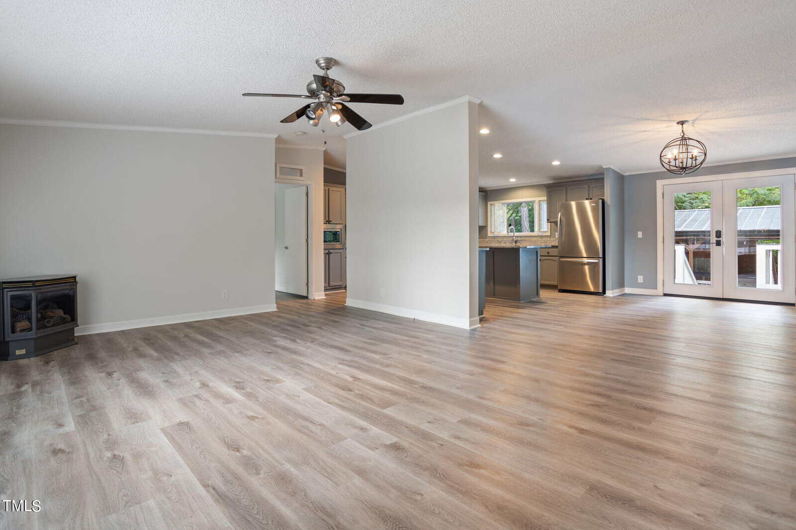 A spacious and bright open-plan living area with light wood flooring. It features a modern kitchen with stainless steel appliances and a stylish light fixture. There's a ceiling fan in the living area and large windows that bring in natural light.