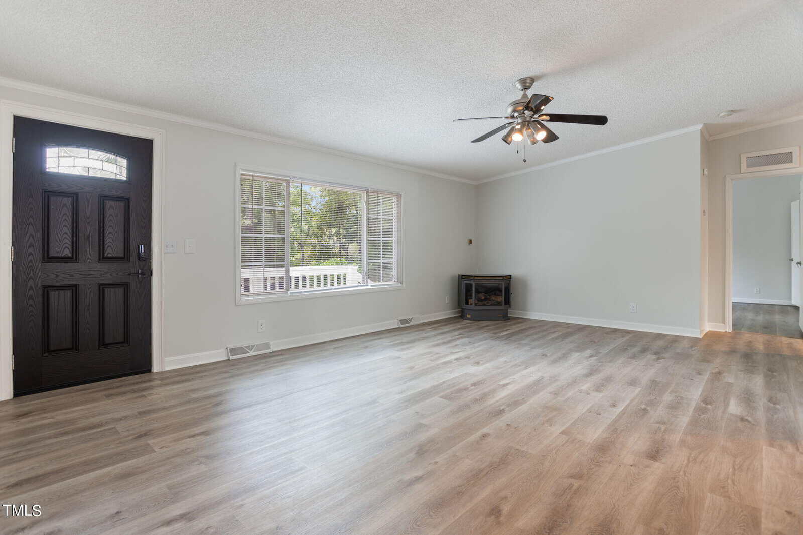 A spacious living room with light wood flooring, neutral-colored walls, a dark wooden front door, and a large window letting in natural light. A ceiling fan with lights is centered on the ceiling, and a small fireplace is in the corner.