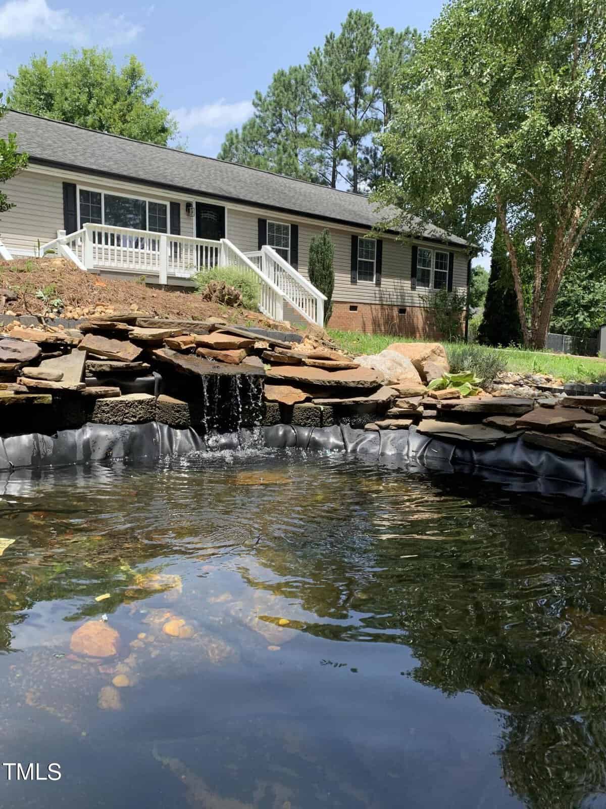 A serene backyard scene with a small waterfall flowing into a pond surrounded by rocks. A single-story house with a porch and railing is visible in the background, framed by green trees and a clear blue sky.