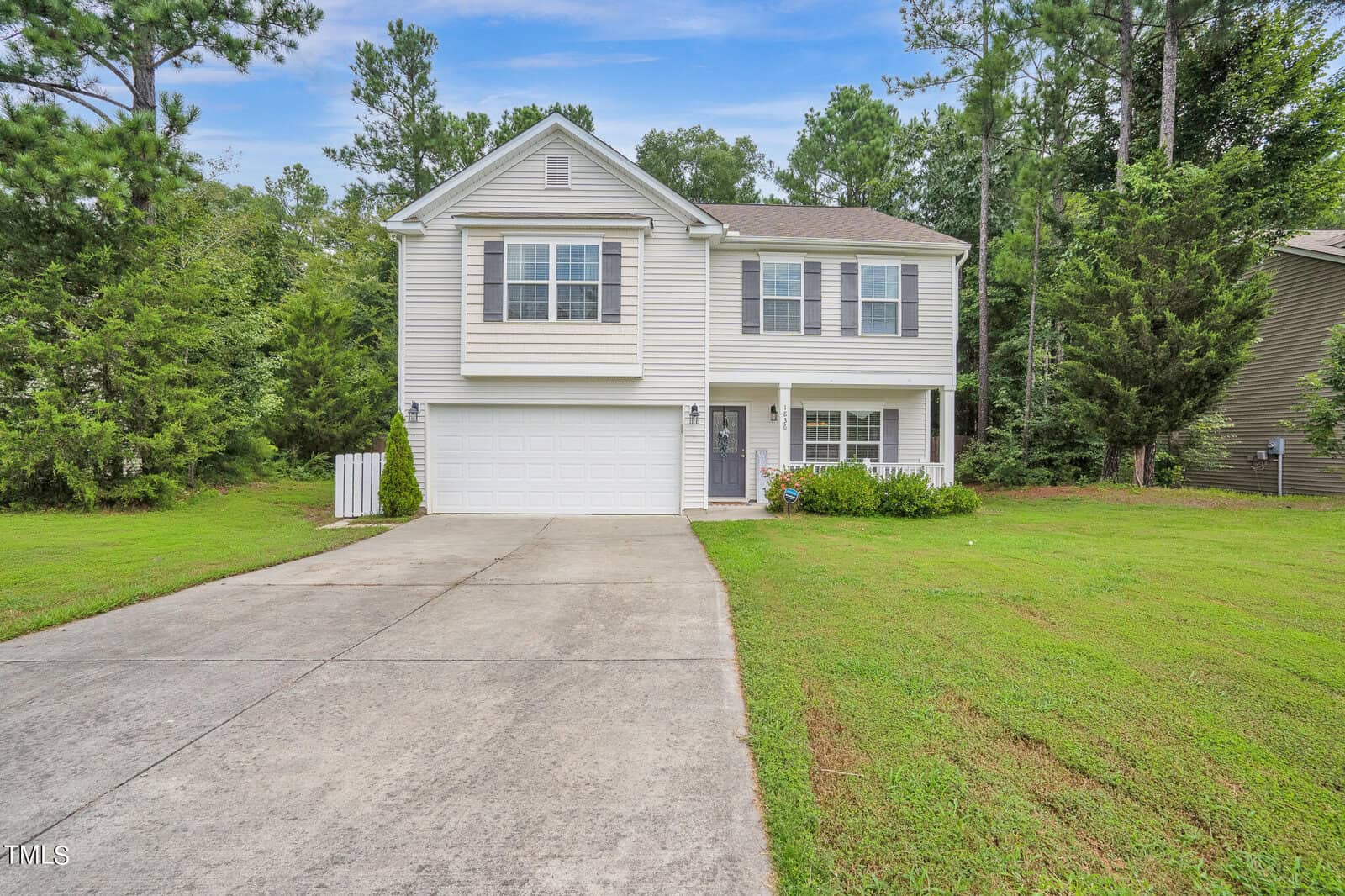A two-story house with a white and gray facade at 1836 Fillmore Drive in Creedmoor is surrounded by trees. It features a double garage, a long driveway, and a neatly trimmed lawn under a clear blue sky.