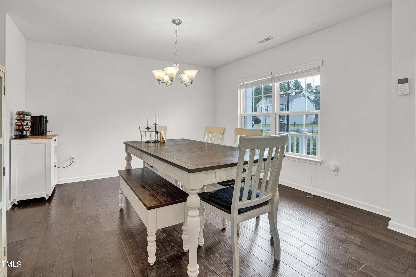 A bright dining room with a wooden table and four chairs. A chandelier hangs above the table, and two candles are placed on it. A window with white blinds offers a view of a house and greenery outside. A small cabinet holds a coffee machine.