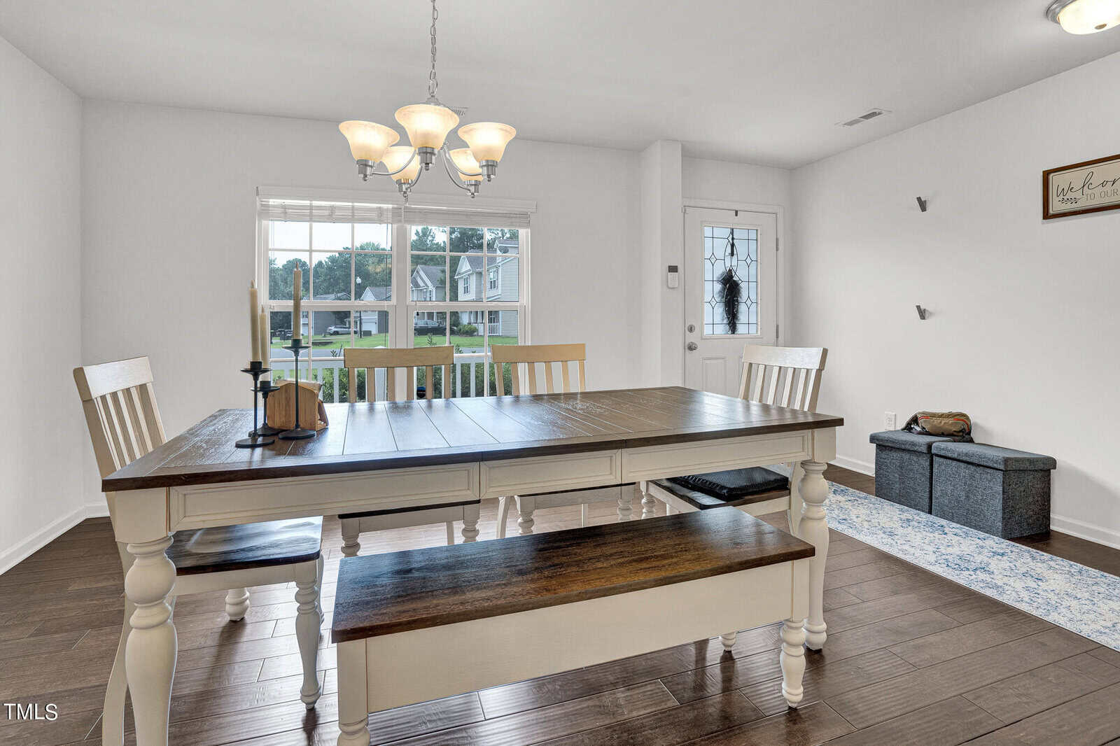 A dining room with a wooden table and matching bench, surrounded by chairs. A chandelier hangs above the table. The room features a large window, hardwood floor, decorative wall sign, and a cushioned storage bench near the door.