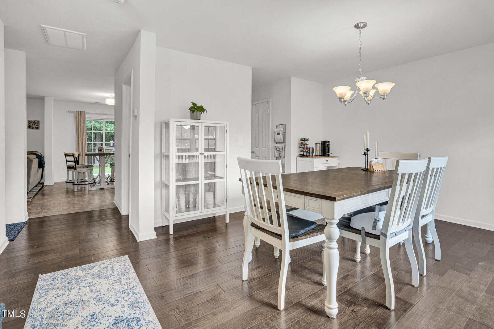 A dining room with white walls and hardwood floors features a white dining table with six chairs. A decorative chandelier hangs above the table. A glass cabinet and small side table are in the background, with natural light from a window.