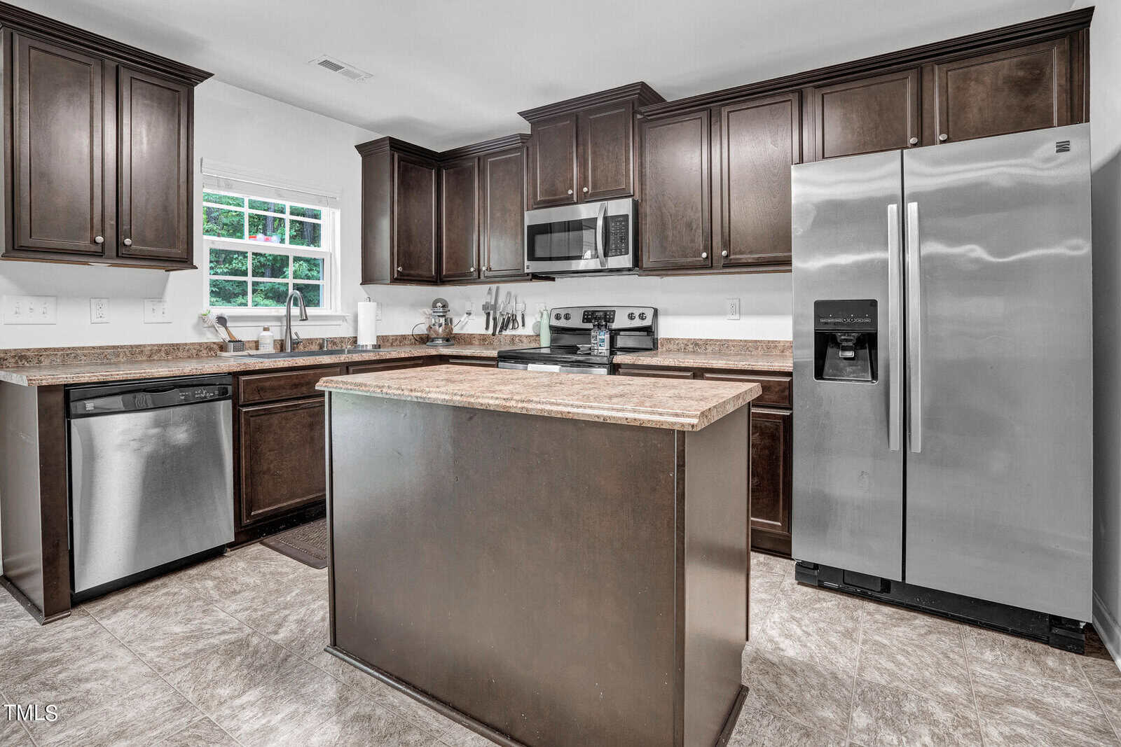 Modern kitchen with dark wood cabinets, stainless steel appliances, a central island with a brown countertop, and tile flooring. There is a window over the sink, and the room has a clean and organized feel.