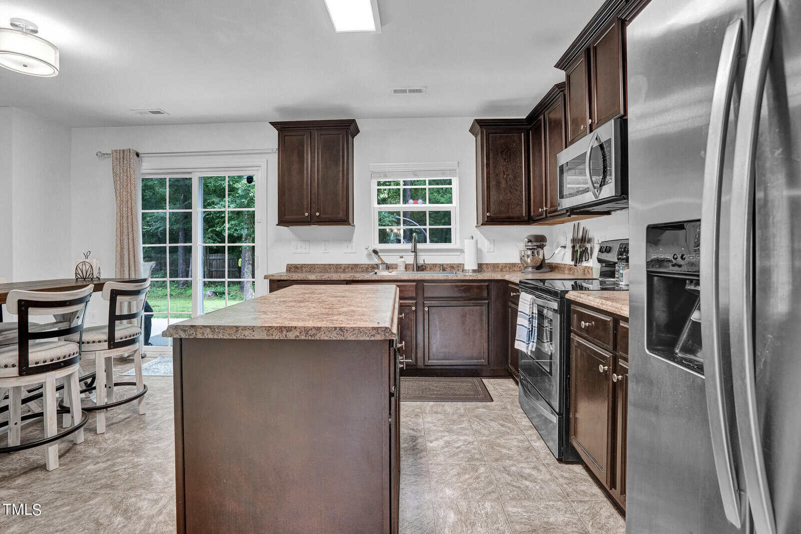 A modern kitchen with dark wood cabinets, stainless steel appliances, and a central island. A dining area with white chairs is visible to the left. Large glass doors offer a view of a green outdoor space.