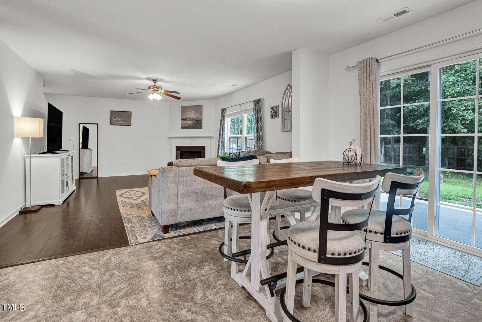 A spacious living room with a modern wooden dining table and four chairs in the foreground. A gray sofa and rug are situated near a fireplace and TV. Large windows with curtains overlook a green backyard, allowing natural light to fill the space.