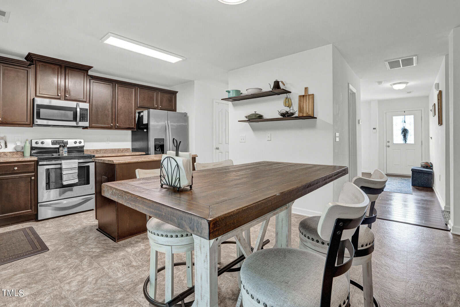 A modern kitchen with dark wood cabinets, stainless steel appliances, and a rustic wooden table with high chairs. The room is bright, with open shelves displaying dishes and decor. A hallway leads to a white front door.