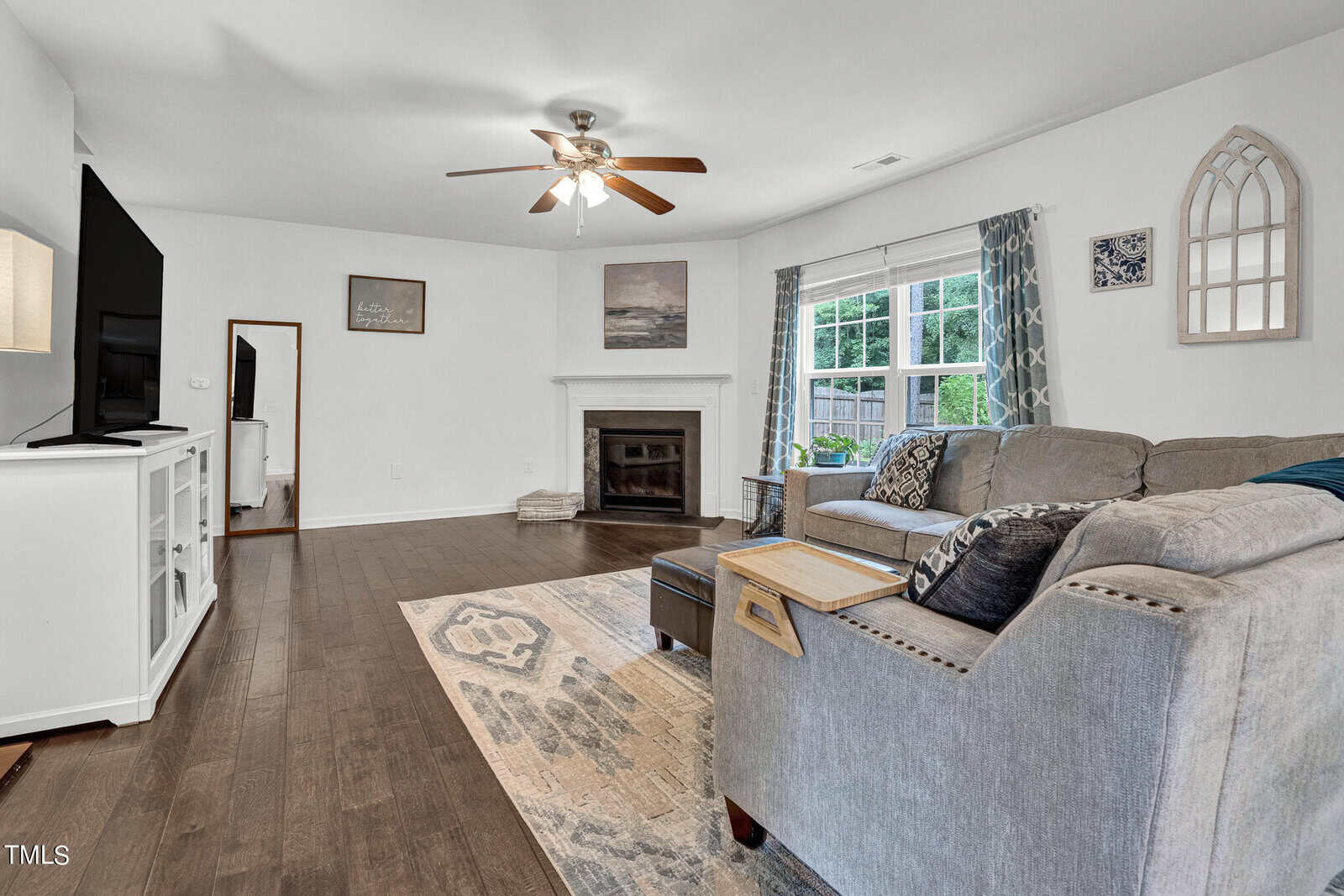A cozy living room with a large gray sectional sofa, a wooden coffee table, and a patterned rug. A flat-screen TV is mounted on a white stand. The room features a fireplace, wall art, and two windows with blue curtains, letting in natural light.