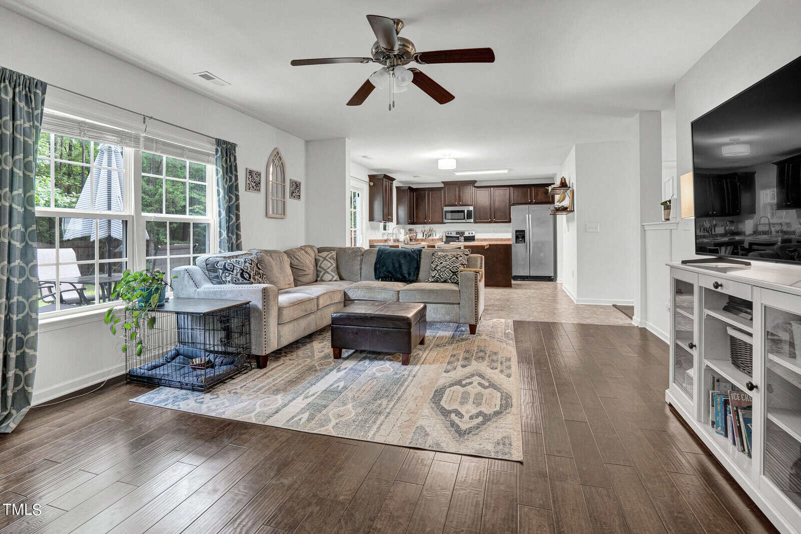 Spacious living room with hardwood floors, a beige sectional sofa, patterned rug, and ceiling fan. A large window provides natural light. The open layout leads to a kitchen with dark cabinets and stainless steel appliances. A TV and dog crate are visible.