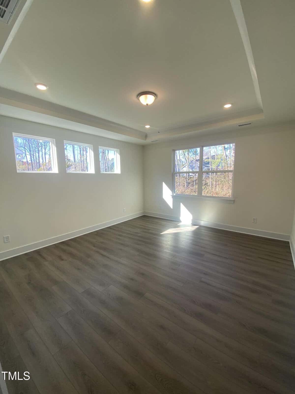 Bright, empty room with light-colored walls, wood flooring, recessed ceiling lights, a central fixture, three small upper windows, and a large window letting in natural sunlight.