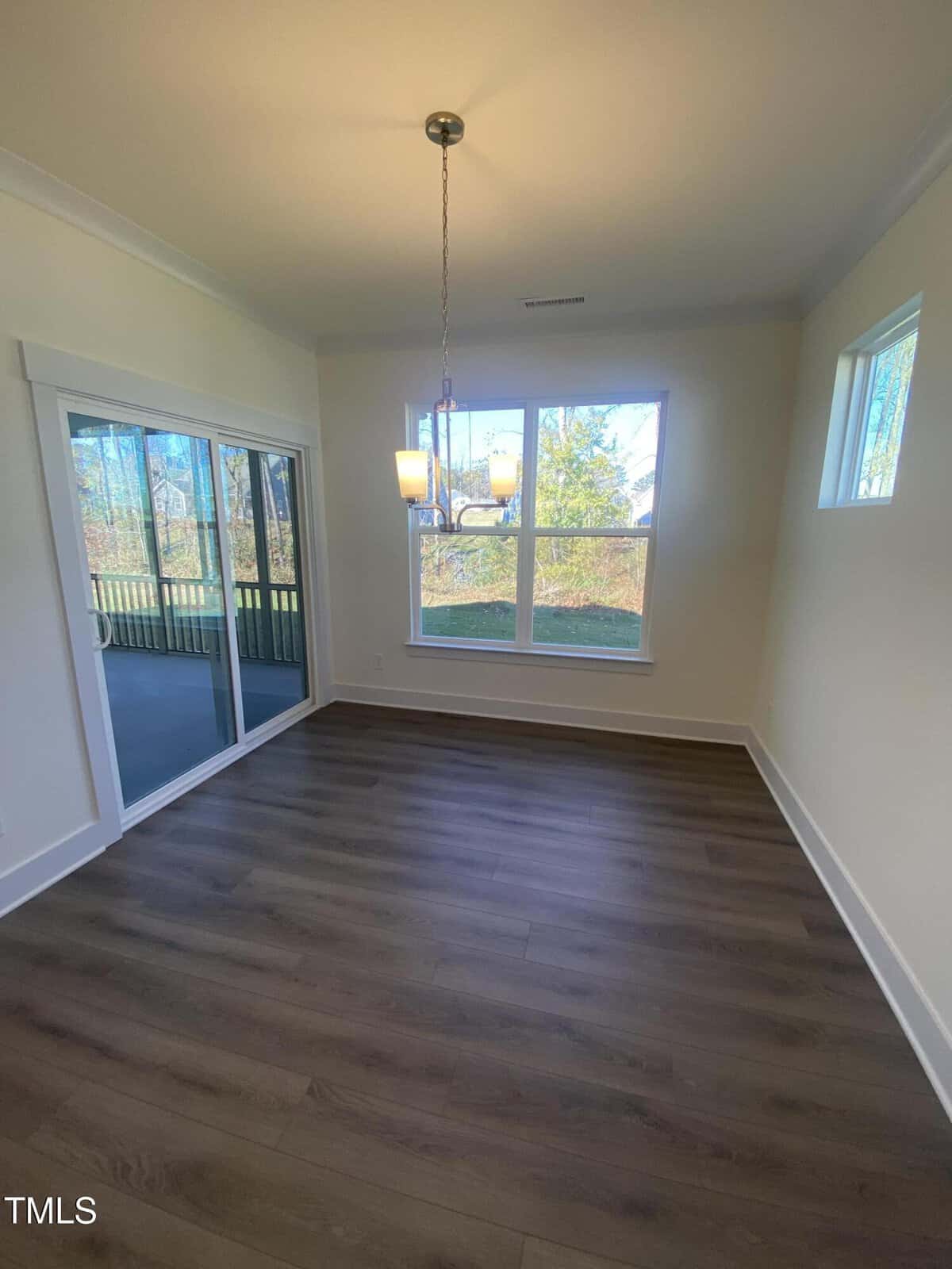 Bright, empty dining area with large windows, a hanging light fixture, wood-style flooring, and sliding glass doors leading to a screened porch. Natural light fills the room, showcasing a view of the outdoors.
