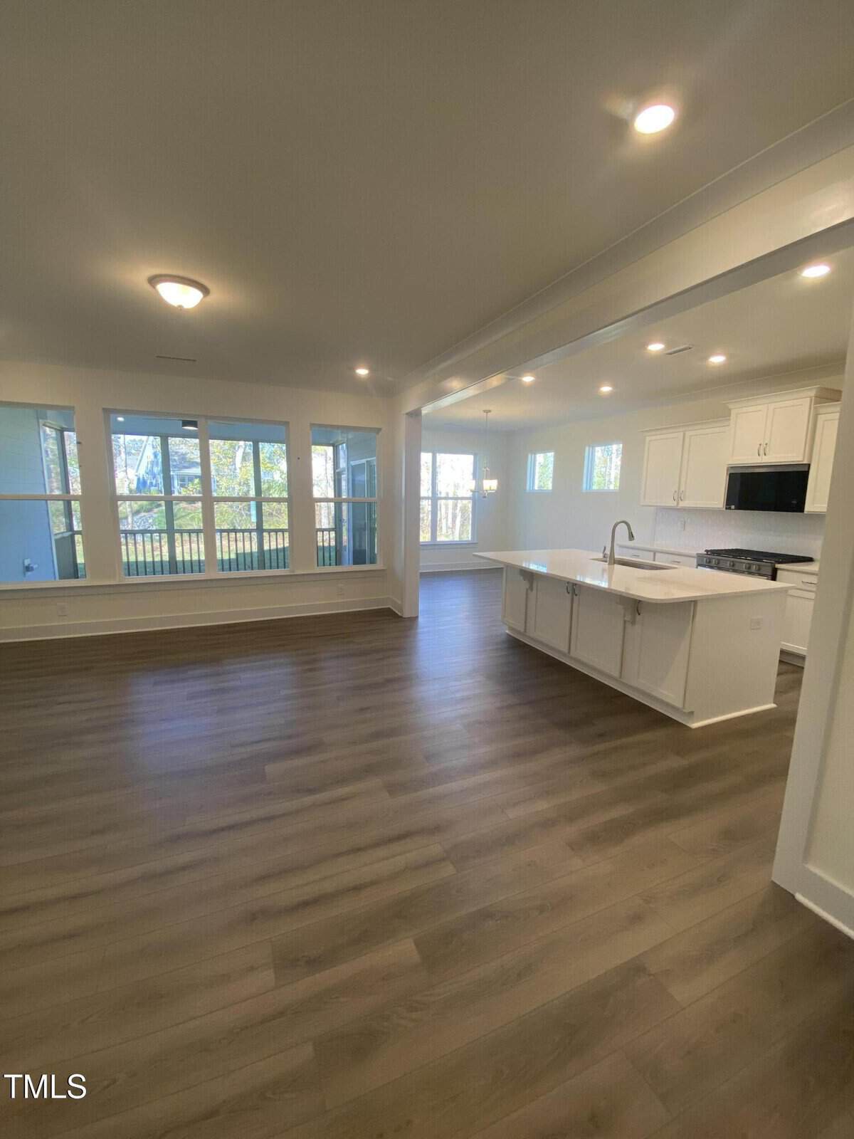 Spacious open-concept living area with wood flooring, large windows, recessed lighting, and a modern kitchen featuring white cabinets, a center island, and stainless steel appliances.