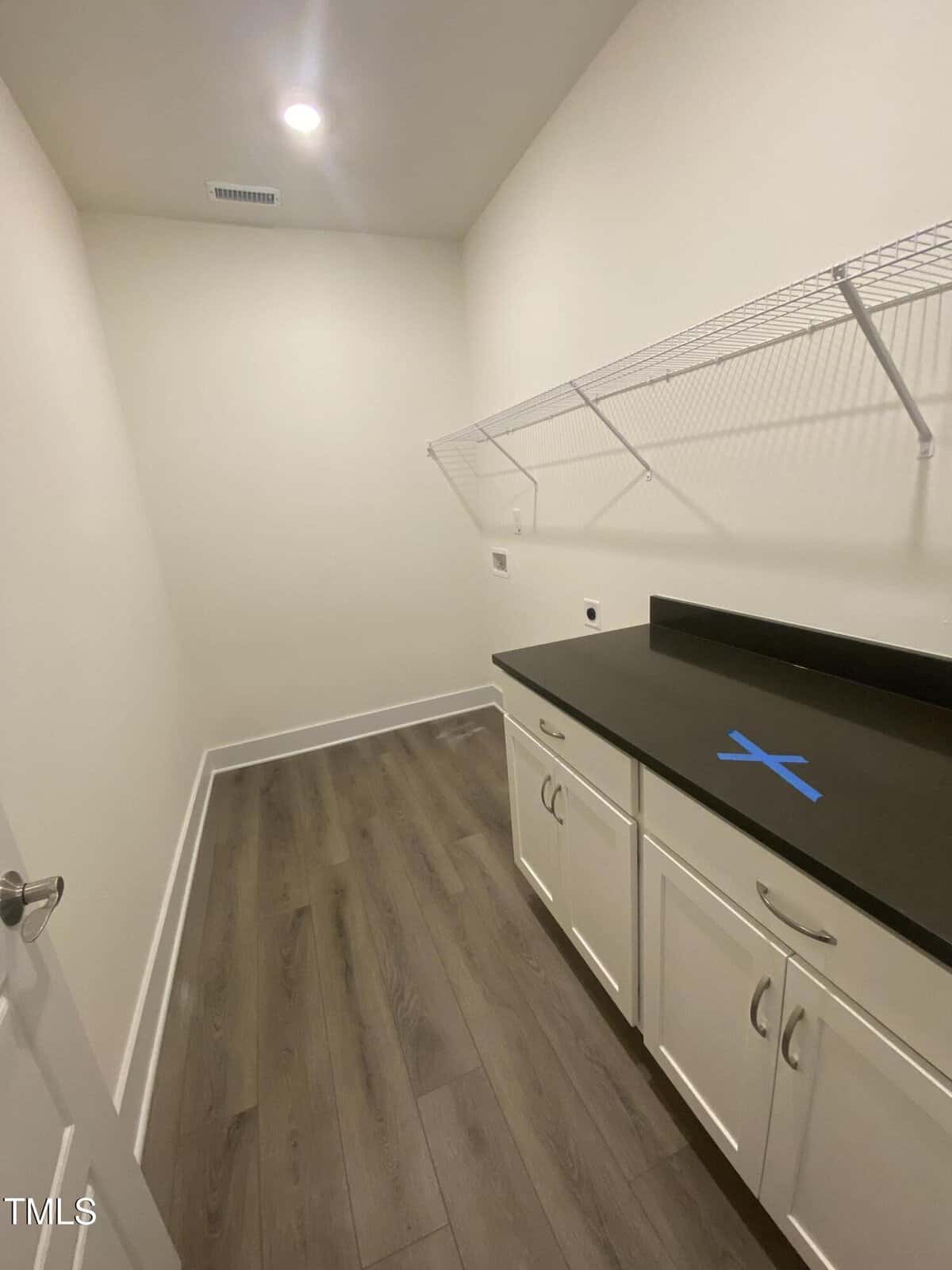 A laundry room with wood flooring, white walls, white cabinets, a black countertop, wire shelf, and hookups for washer and dryer. A blue tape “X” is on the counter. Ceiling light is on.