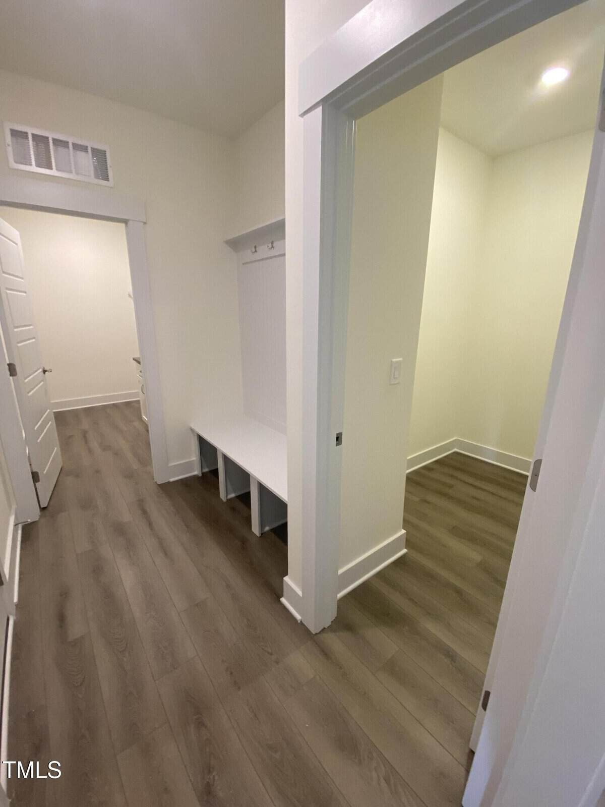 A hallway with light wood flooring, white walls, and white trim; a built-in bench with cubbies and hooks is on the left, and an open doorway leads to a small, well-lit room on the right.