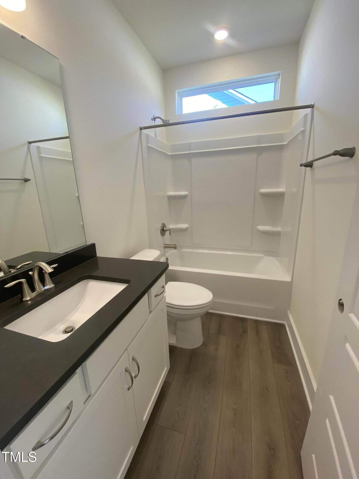 Bright, modern bathroom with white walls, a bathtub-shower combo, white vanity with black countertop and silver faucet, toilet, large mirror, and wood-style flooring. Natural light enters through a small window above the tub.