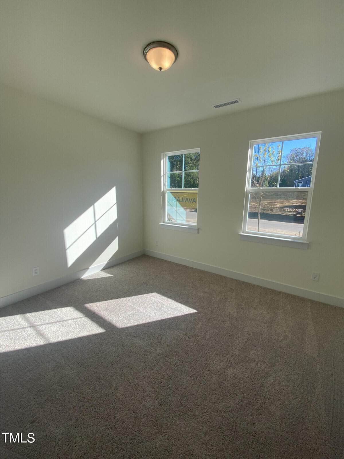 A bright, empty room with beige carpet, white walls, and two large windows letting in natural light. Sunlight casts clear shadows on the floor and wall. A ceiling light fixture is centered overhead.