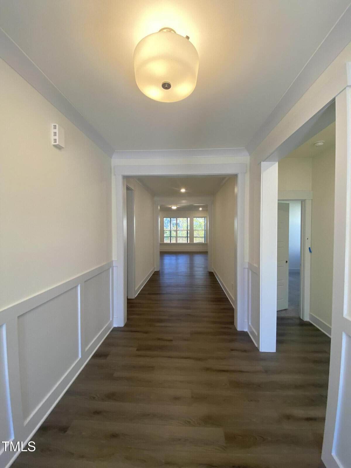 A hallway with light-colored walls, wood flooring, white trim, and a ceiling light fixture leads to a bright room with large windows and a view of trees outside. Rooms or doorways are visible on both sides of the hallway.