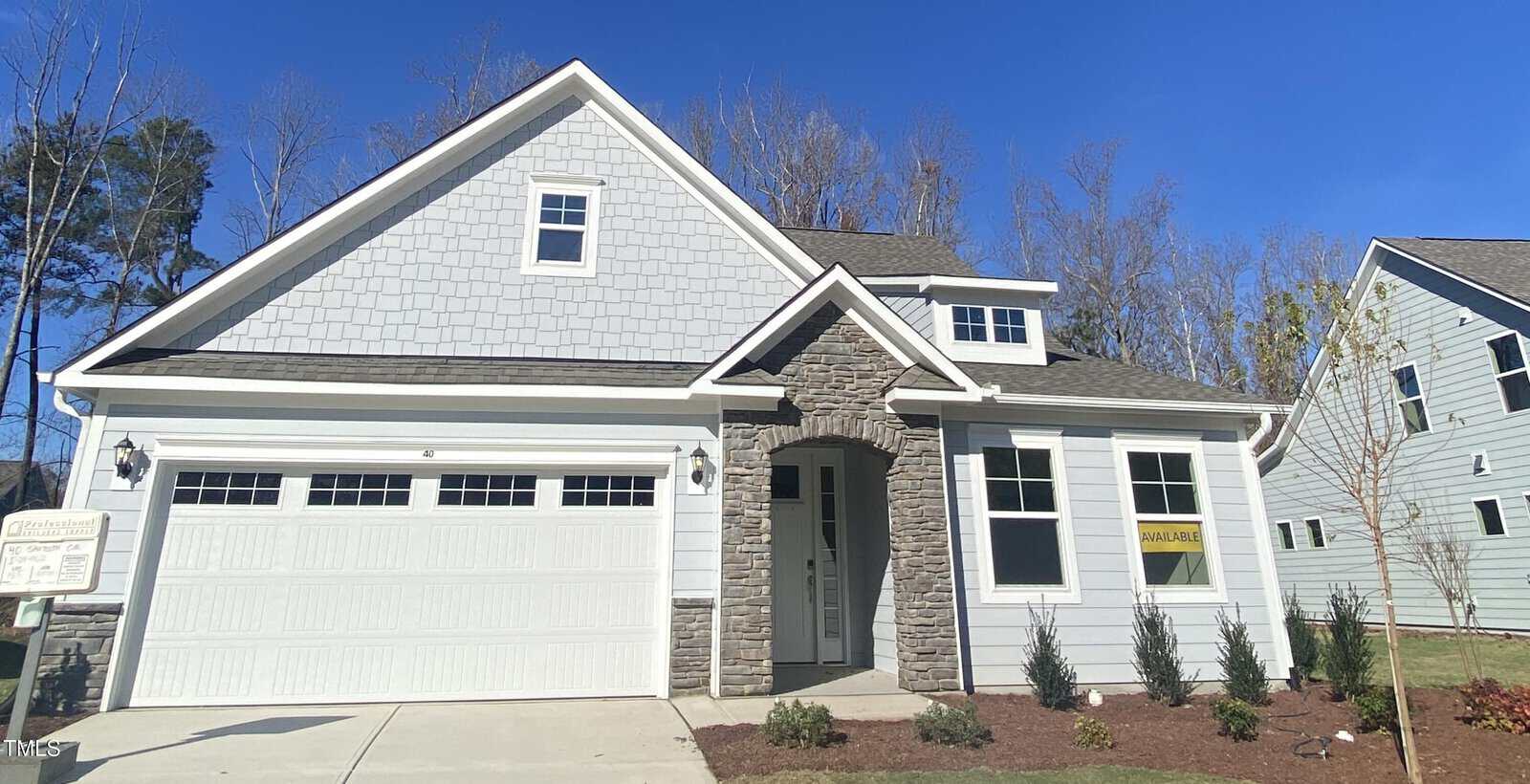 A modern, two-story house with light gray siding, white trim, a double garage, and a stone arch entryway. There are small shrubs in the front yard and a "for sale" sign in a window. The sky is clear and blue.