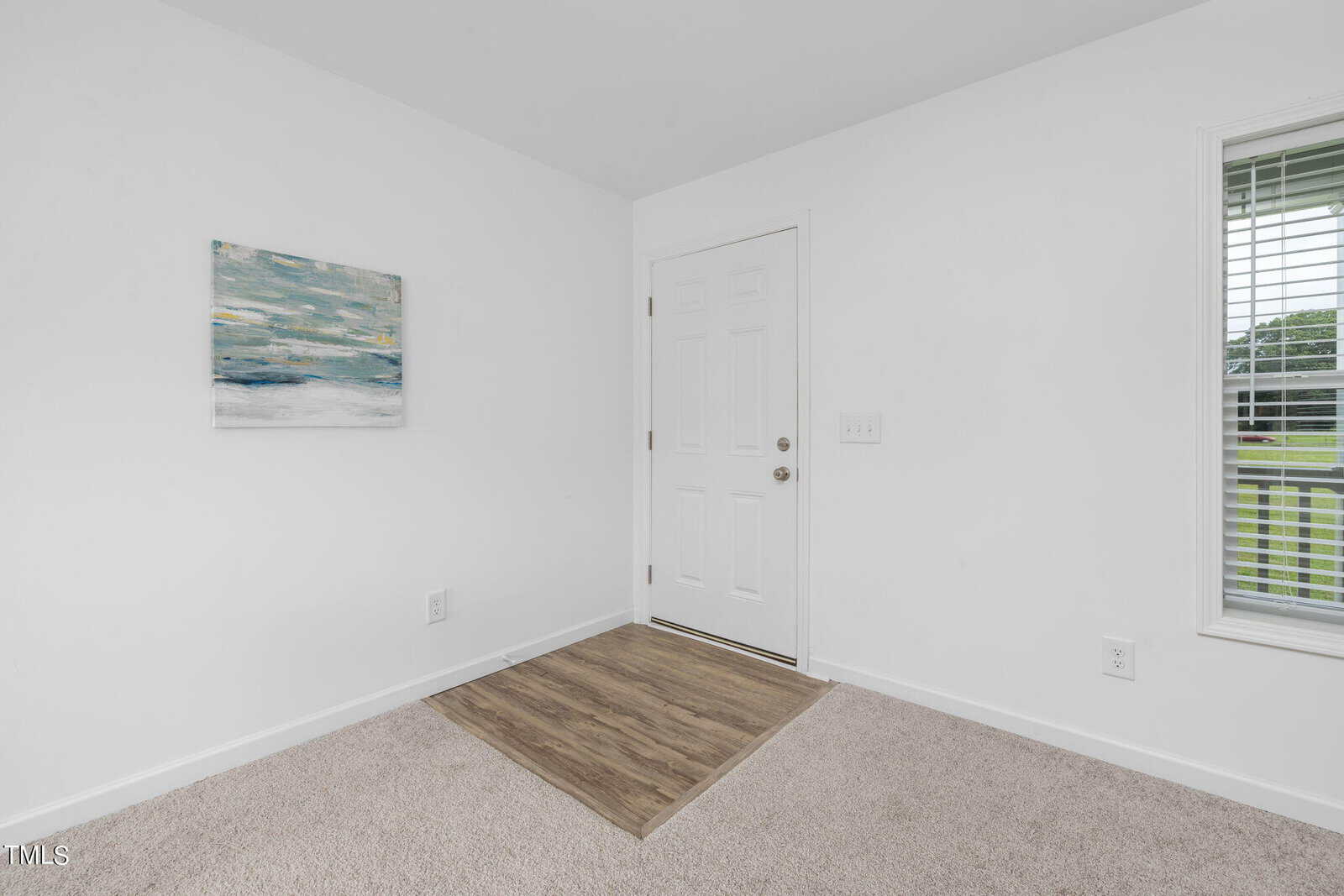 A minimalist room with white walls, a door with a doorknob at the center of the wall, and a window with blinds to the right. The floor is carpeted, except for a small section of wood-look flooring in front of the door. A seascape painting hangs to the left of the door.