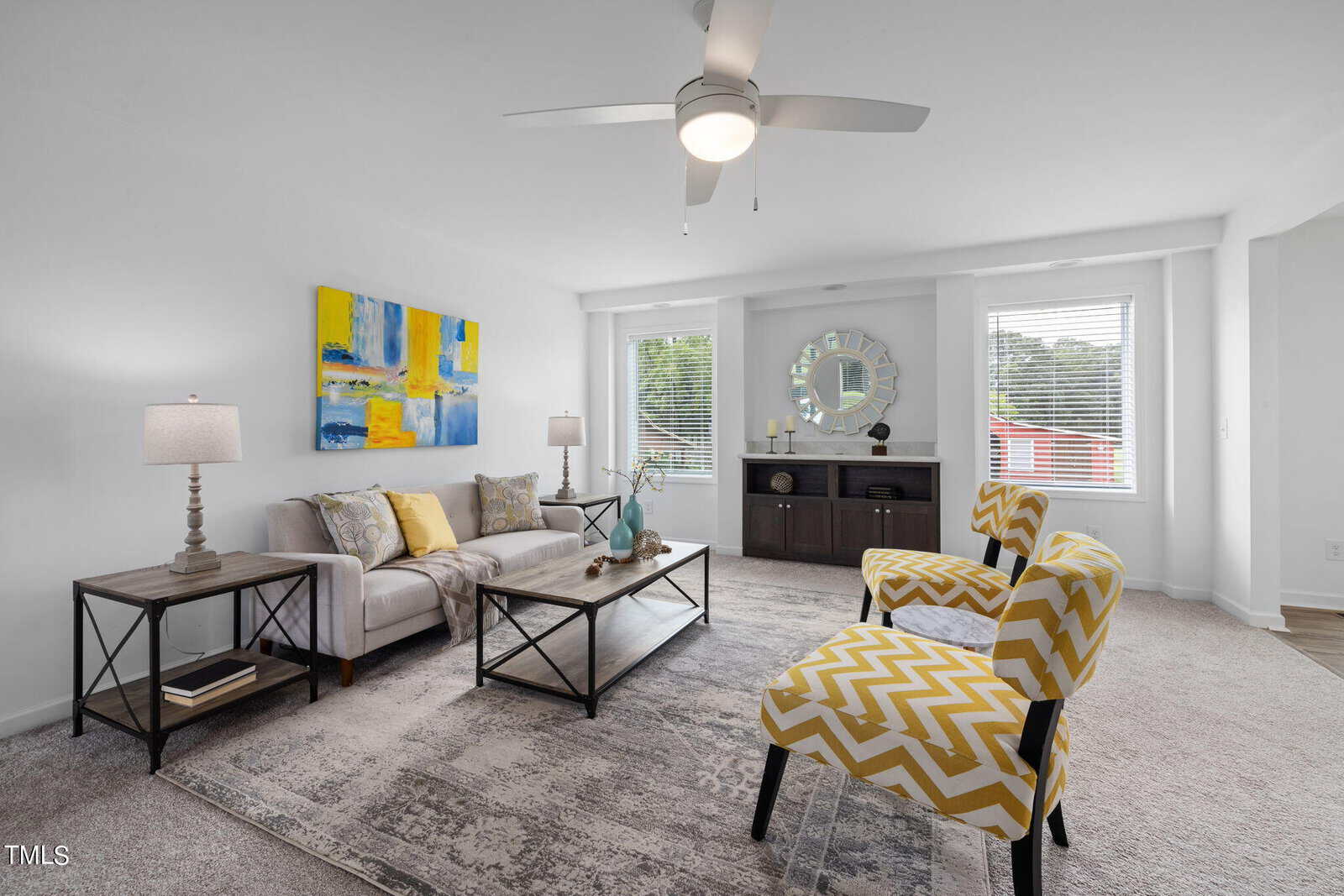 A modern living room with a gray couch, colorful abstract art above it, and two bright yellow zigzag-patterned armchairs. A coffee table and side tables with lamps are present. A large window with blinds brings in natural light. The room is decorated in neutral tones.