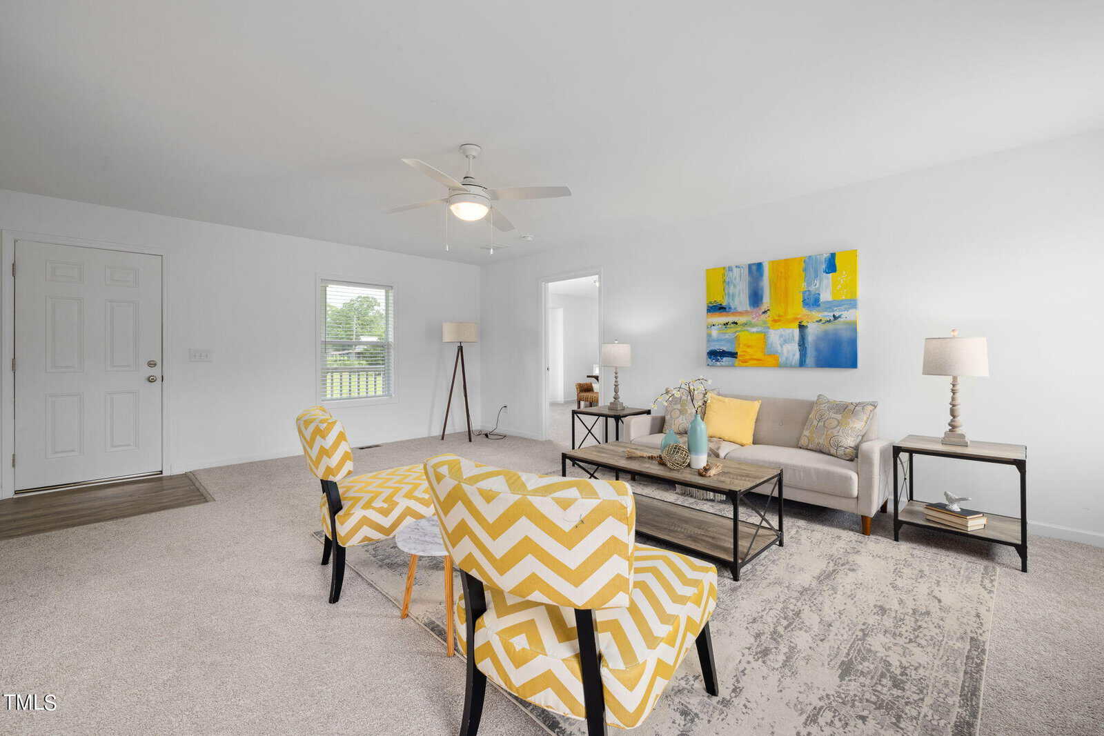 A modern living room with yellow zigzag patterned chairs, a beige sofa with decorative pillows, and a wooden coffee table. The walls are white, with a colorful abstract painting above the sofa. Lamps are placed beside the sofa and near the window. A ceiling fan is overhead.