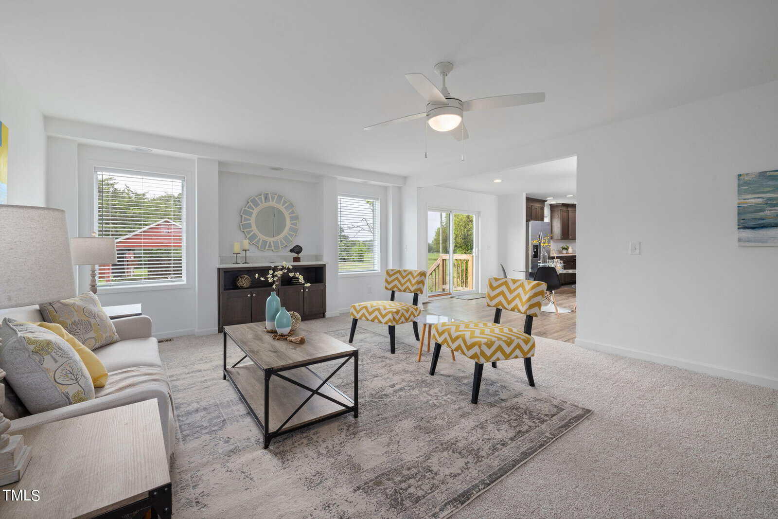 A bright living room with a beige couch, yellow and white patterned chairs, and a wooden coffee table on a light area rug. Large windows bring in natural light, and a ceiling fan hangs above. In the background, there's an open doorway leading to a kitchen.