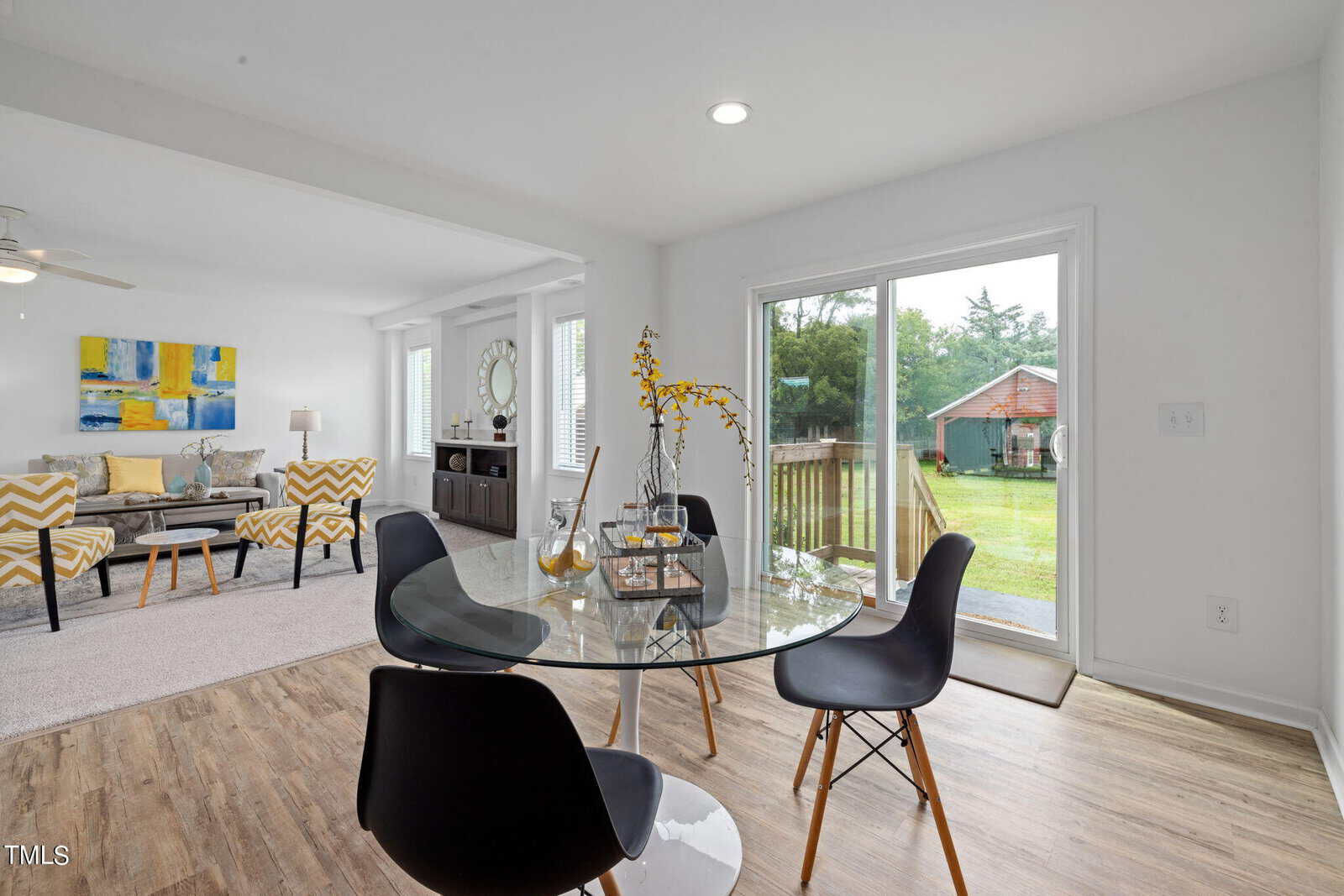 A modern dining area with a glass table and four black chairs, positioned next to a sliding glass door that opens to a backyard. A living space with a gray sofa, yellow and gray-patterned chairs, and a colorful abstract painting is visible in the background.