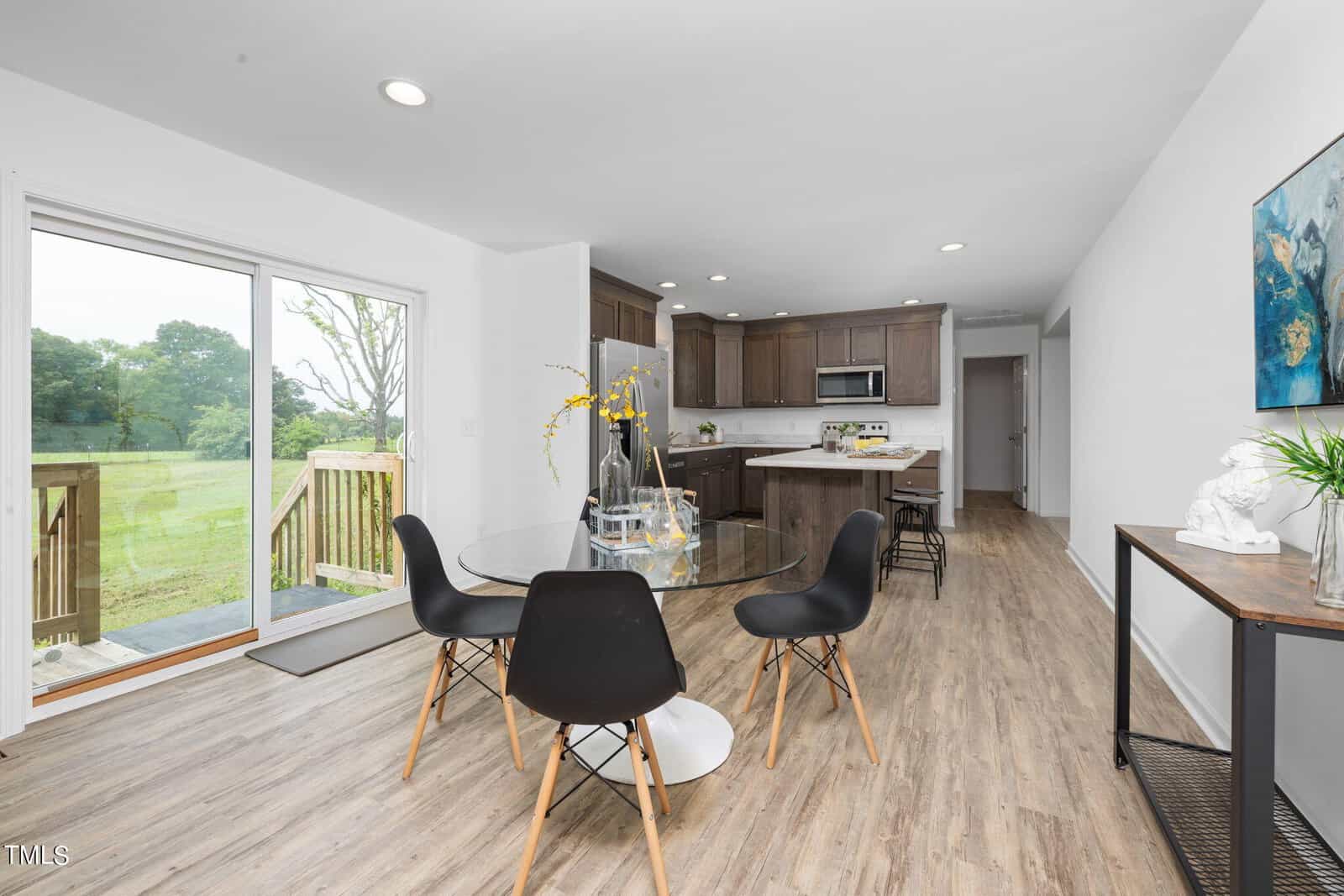 A modern, open-concept dining and kitchen area. The dining space features a round glass table with four black chairs. Behind, the kitchen showcases dark wood cabinets and stainless steel appliances. A large sliding door opens to a deck with a view of greenery outside.