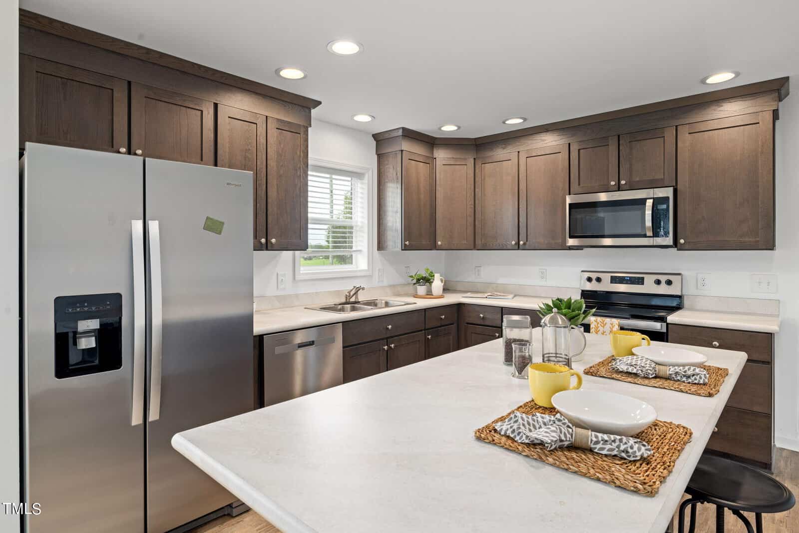 Modern kitchen with dark wood cabinets, stainless steel refrigerator, oven, microwave, and dishwasher. An island in the foreground has two place settings with white bowls and yellow mugs on woven placemats. A window above the sink lets in natural light.