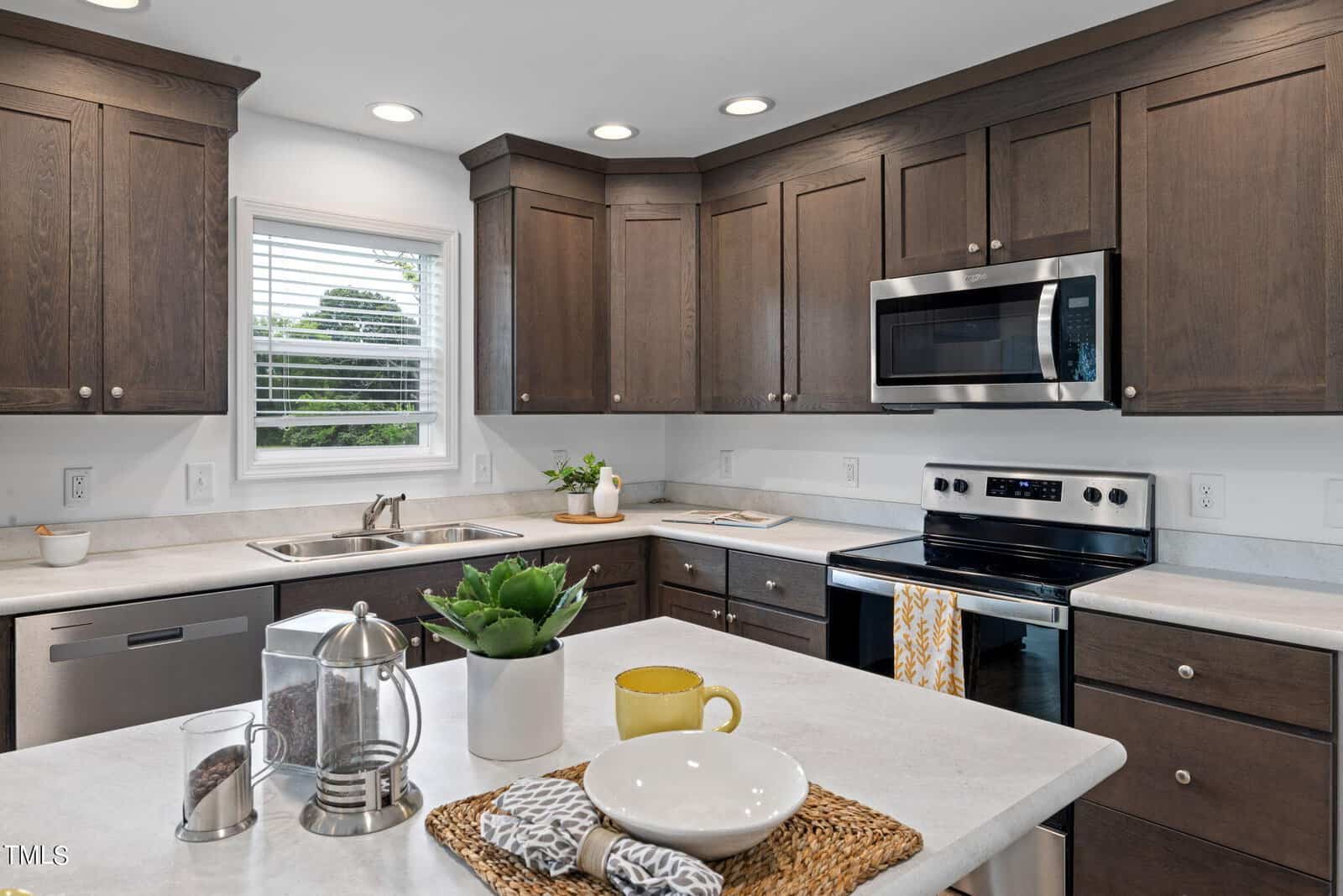 A modern kitchen with dark wooden cabinets, white countertops, and stainless steel appliances, including a built-in oven and microwave. A kitchen island holds a potted plant, coffee pot, yellow mug, and a woven tray holding a tea towel. A window with blinds is above the sink.