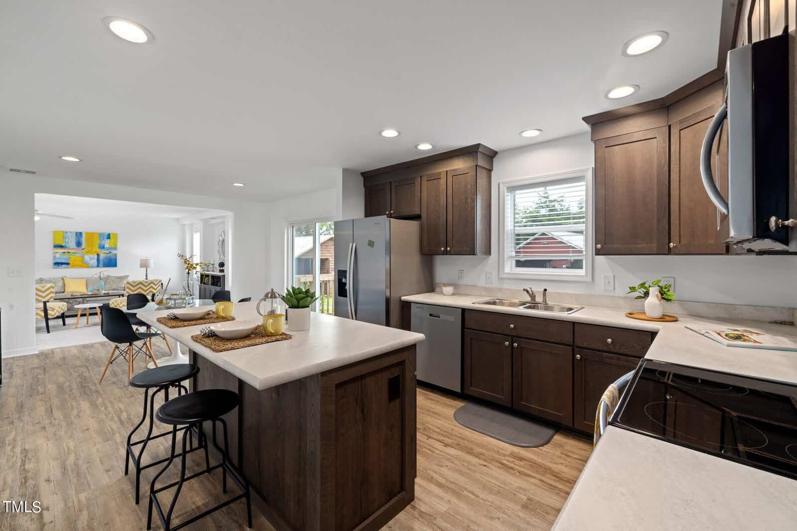 Modern kitchen interior with dark wood cabinetry and white countertops. An island with barstools is on the left, and stainless steel appliances are featured. The space opens up to a bright dining and living area with large windows, light wood flooring, and contemporary decor.
