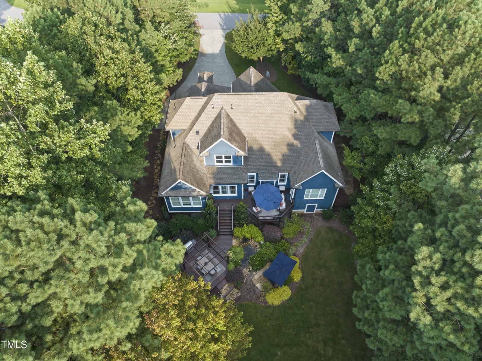 Aerial view of a large house surrounded by trees. The property features a stone driveway, a backyard with a circular patio area, and landscaped gardens. The house has a gabled roof and dark exterior siding.